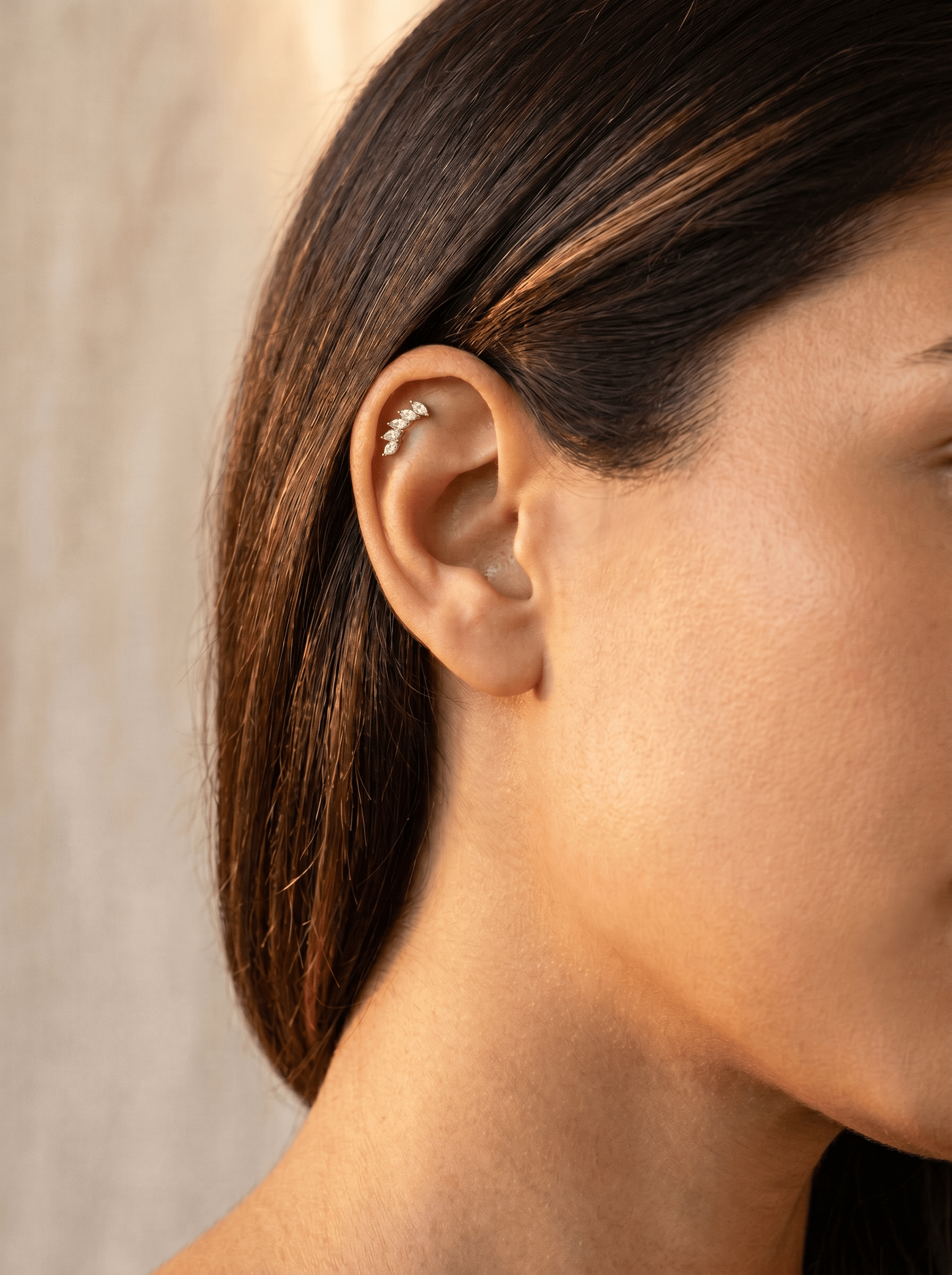 Primer plano de la oreja de una mujer adornada con el pendiente tipo «ear cuff» «Gia dorado» de Titanio-Noble Art. El diseño de rama con hojas realza su look, que luce sobre una piel de tonos cálidos y con el pelo castaño recogido hacia atrás, todo ello sin necesidad de perforación. El fondo aparece ligeramente desenfocado.