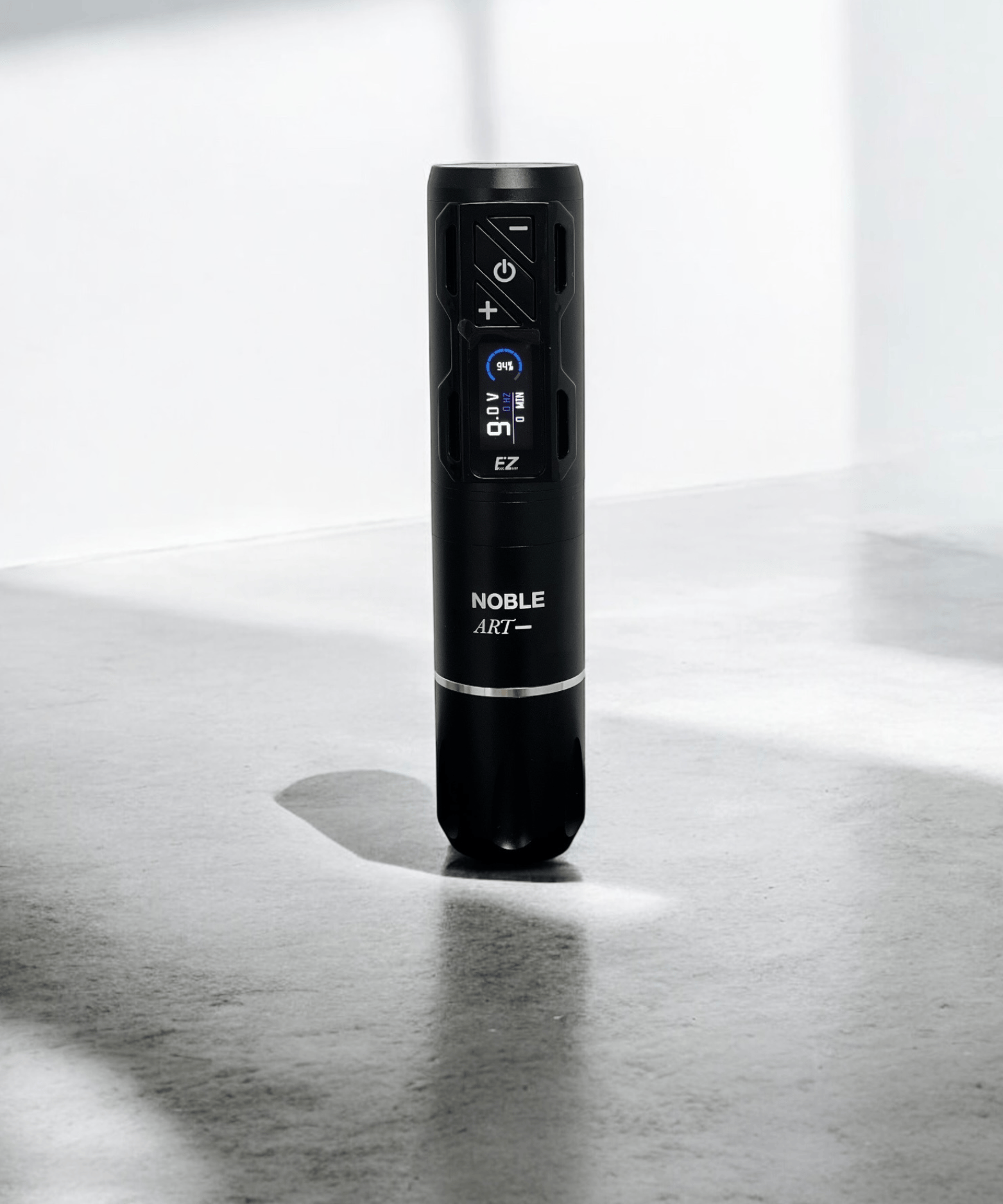 Una máquina de tatuar rotativa inalámbrica EZ 2PRO Wireless de Noble Art, de color negro y forma cilíndrica, se encuentra en posición vertical sobre una superficie gris con sombras marcadas y una luz suave; cuenta con una pantalla digital y botones de control para tatuar o hacer piercings.