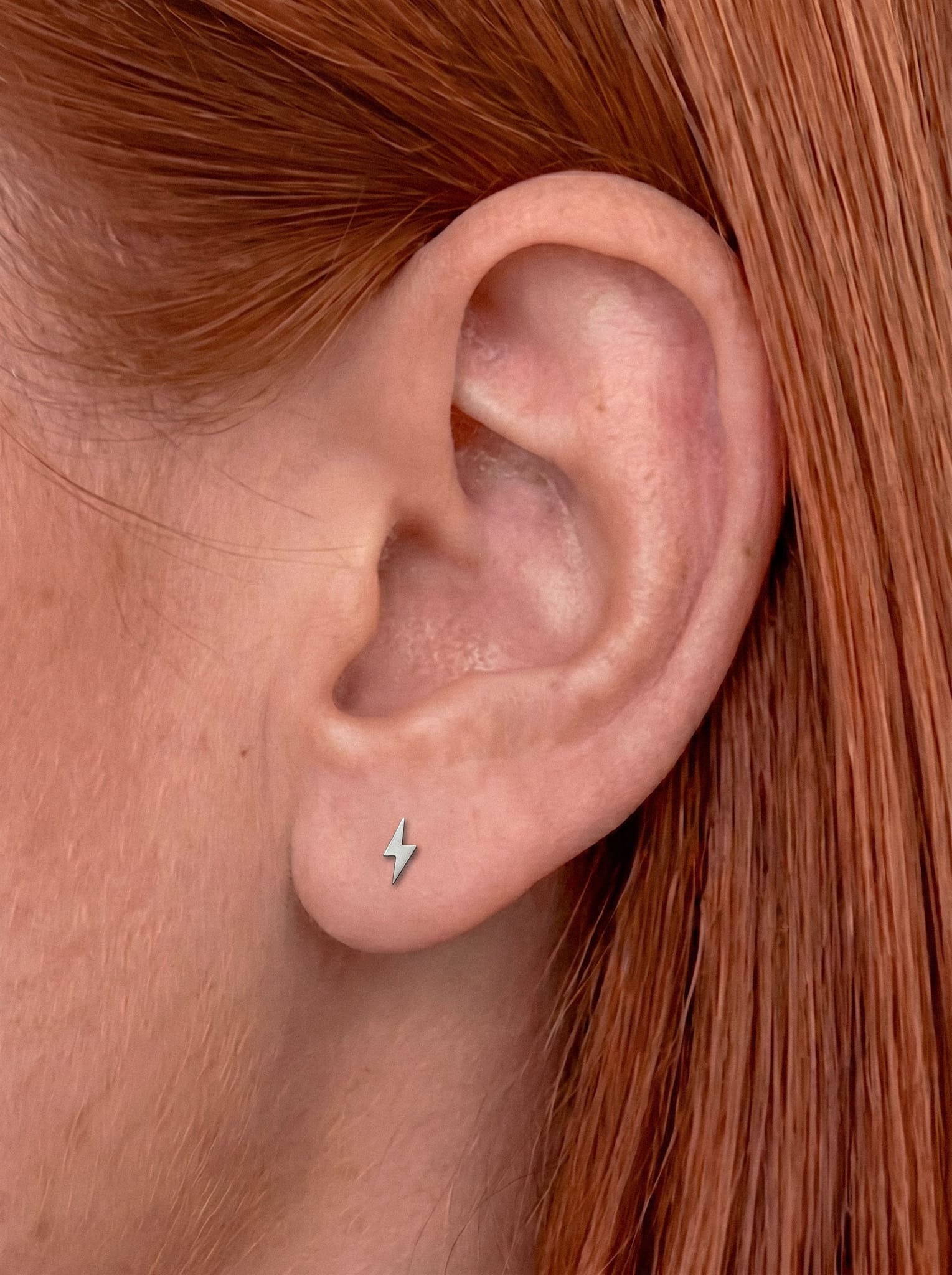 Primer plano de la oreja de una persona con el pelo liso y rojo y un piercing, que lleva el pendiente de plata «Rayo liso», con forma de rayo, de la marca Titanio-Noble Art.
