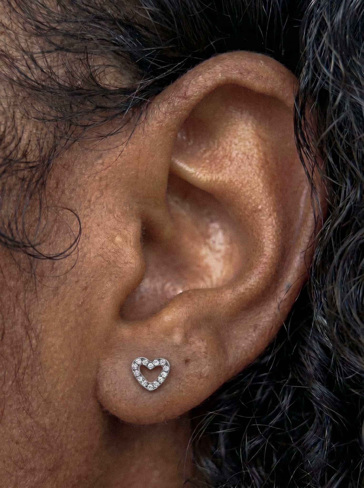 En un primer plano se aprecia un pendiente tipo stud con forma de corazón de la colección «Love liso» de Titanio-Noble Art, adornado con pequeñas piedras preciosas. Un cabello oscuro y rizado enmarca la oreja, resaltando la elegancia de este accesorio.