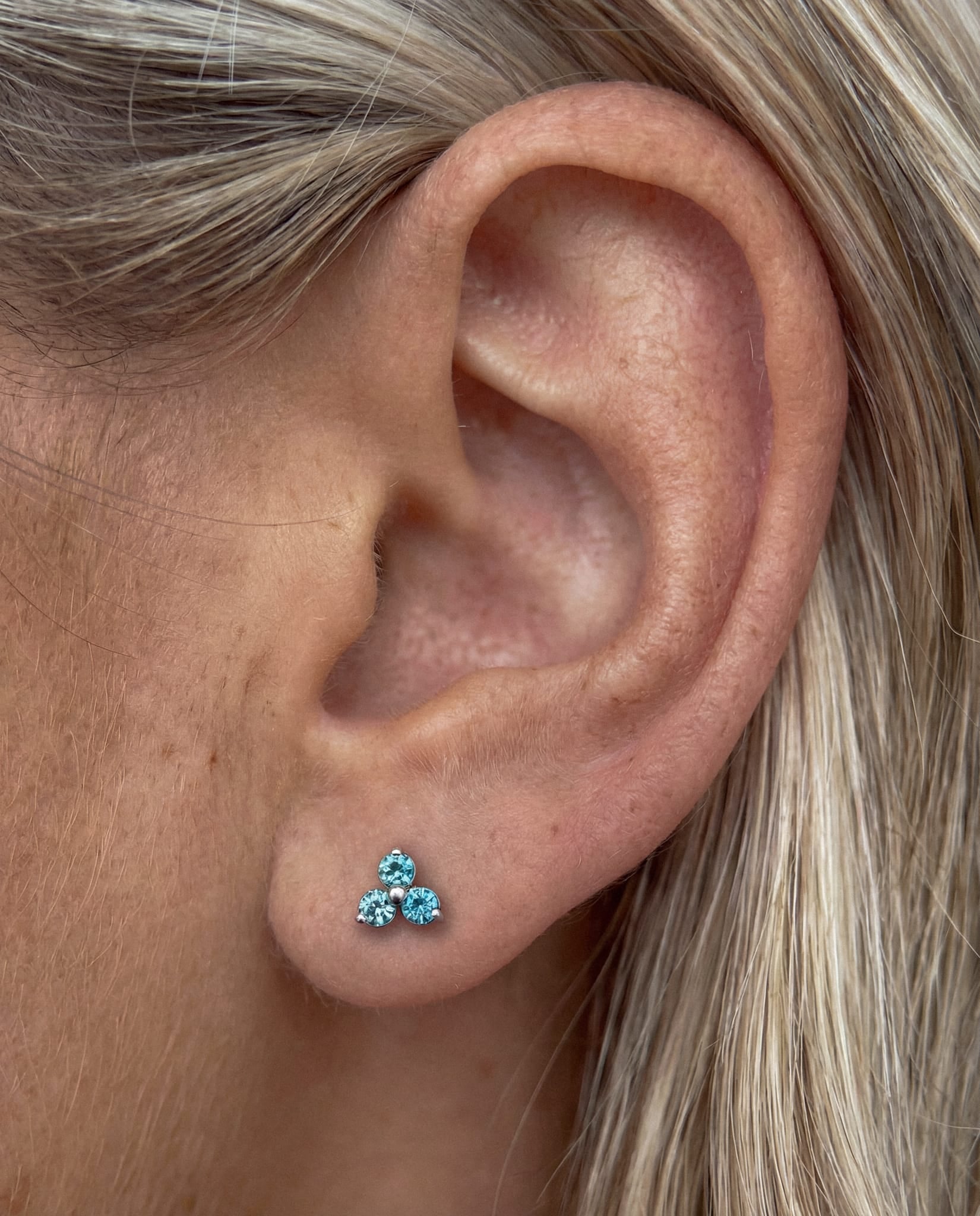 Primer plano de la oreja de una persona con el pelo rubio claro, que lleva el modelo «Trinity cz» de Titanio-Noble Art: un pequeño pendiente de botón con tres piedras preciosas azules dispuestas en forma de triángulo, lo que resalta su elegancia.