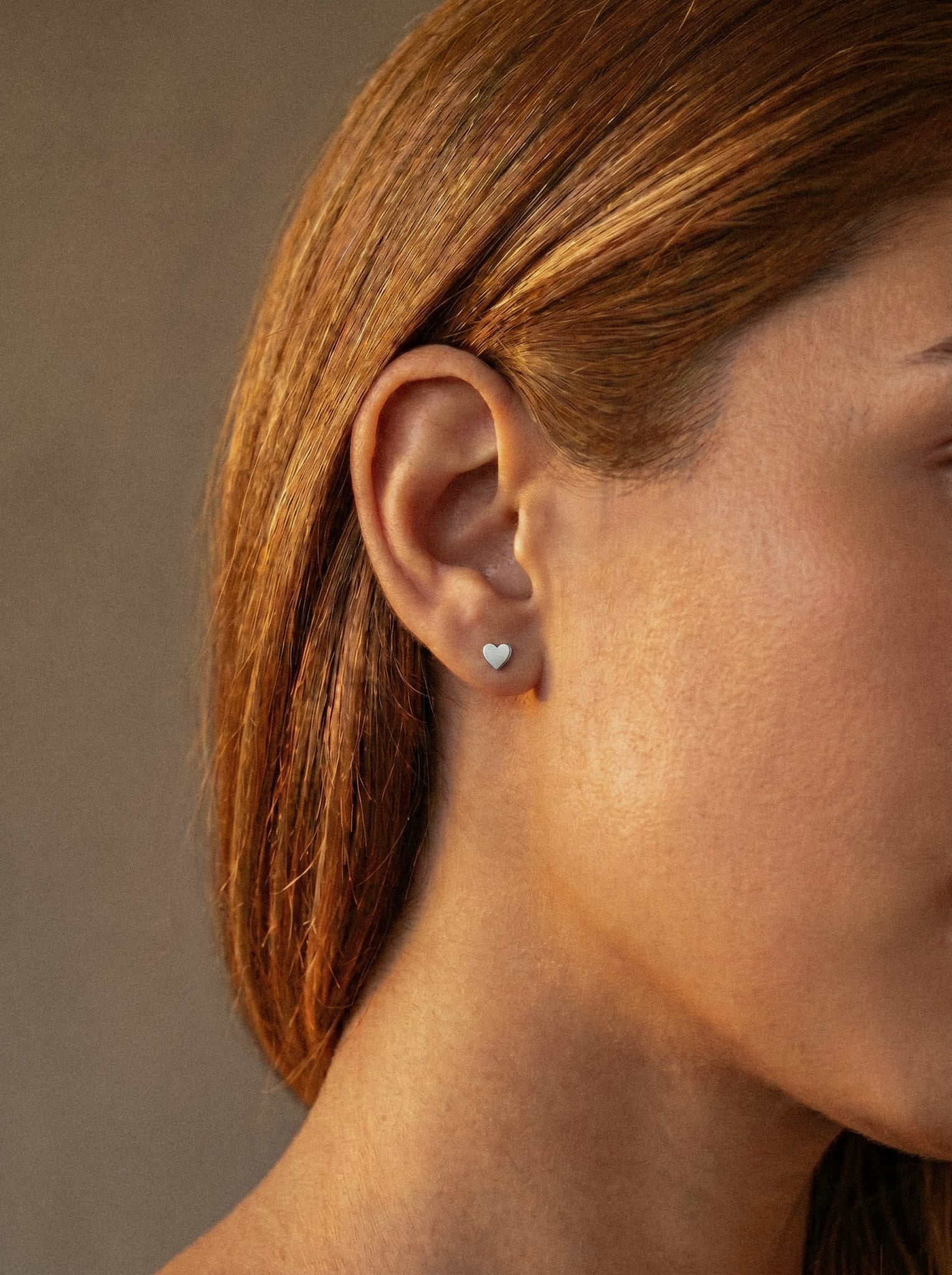 Primer plano de una melena lisa de color castaño claro que enmarca un pequeño pendiente de botón en forma de corazón «LOVE LISO SILVER», de la marca Titanio-Noble Art, en la oreja izquierda. Un tatuaje descolorido cerca de la oreja sugiere que se ha sometido recientemente a una eliminación con láser. El fondo, suavemente iluminado, es de color neutro.
