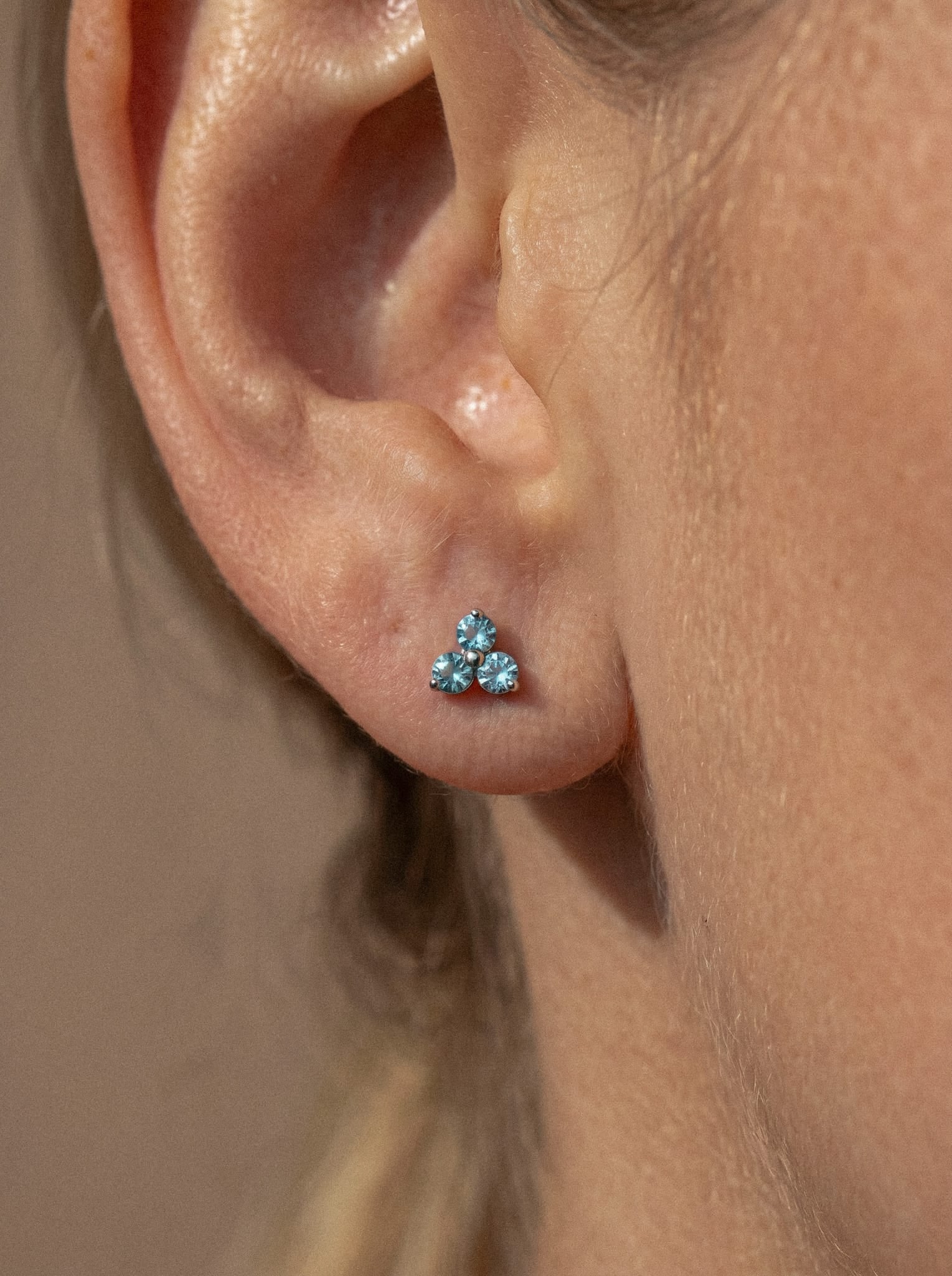 Primer plano de la oreja de una persona que lleva el pendiente «Trinity cz» de Titanio-Noble Art, con tres piedras preciosas de color azul claro dispuestas en forma de triángulo, lo que pone de relieve la exquisita artesanía joyera.
