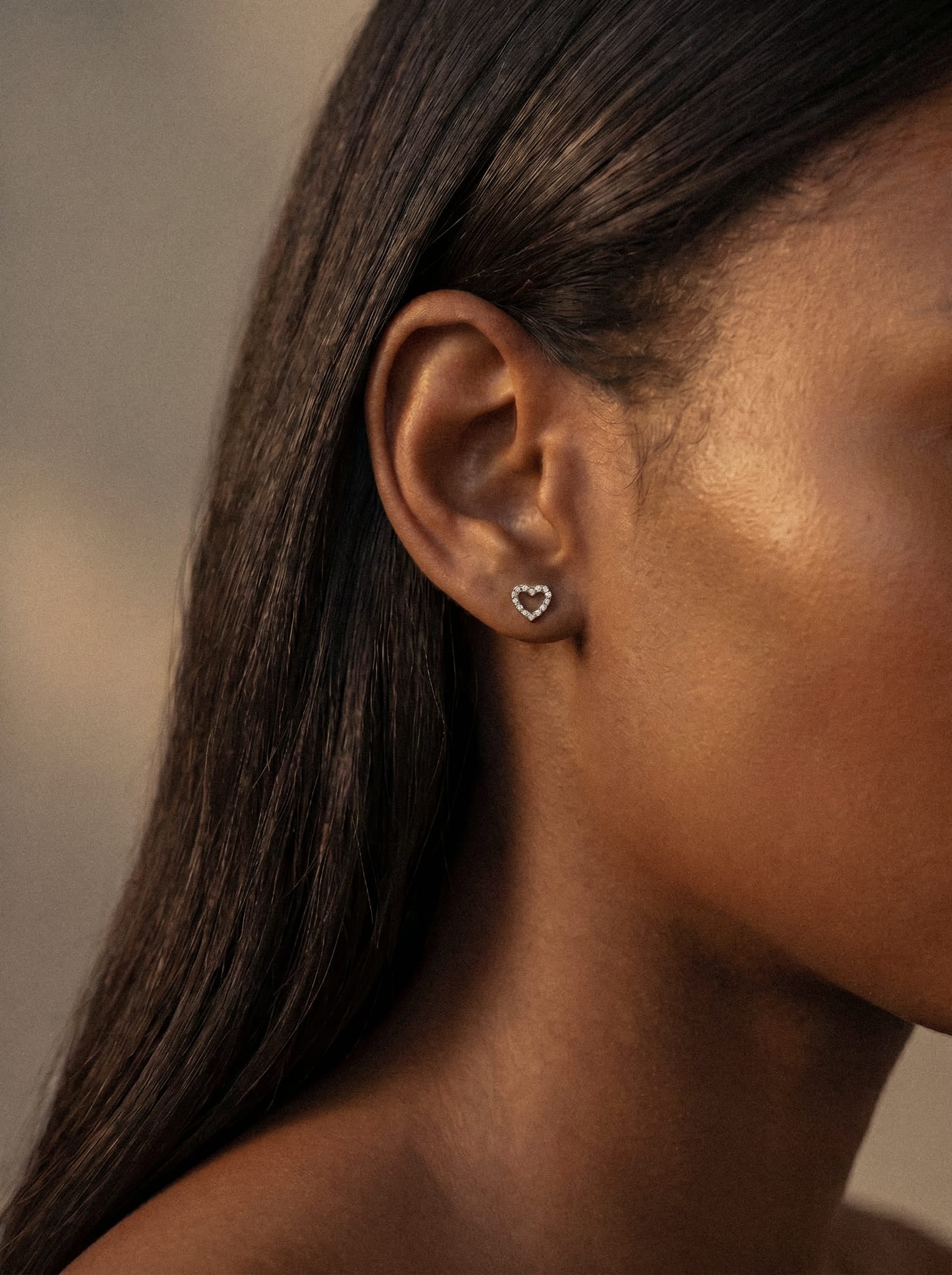 Primer plano de la oreja de una mujer que lleva el pendiente de botón «Love liso» con forma de corazón de Titanio - Noble Art. Su pelo liso y oscuro está recogido detrás de la oreja, y su piel cálida y radiante contrasta con un fondo suavemente difuminado.