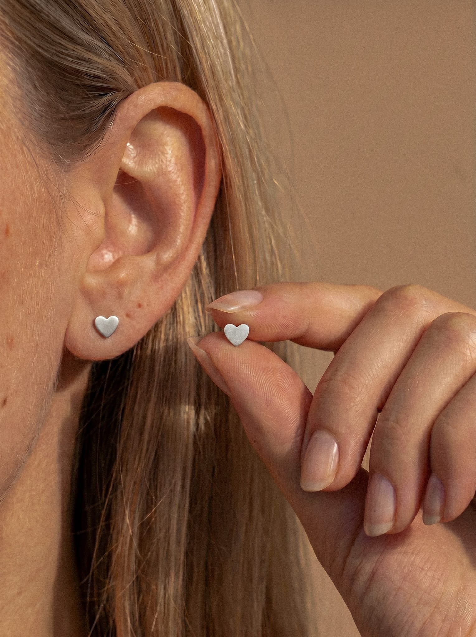 En un primer plano se ve a una persona rubia que lleva el pendiente de corazón «LOVE LISO SILVER» de Titanio-Noble Art, sosteniendo el pendiente a juego. El elegante look destaca sobre un fondo neutro.