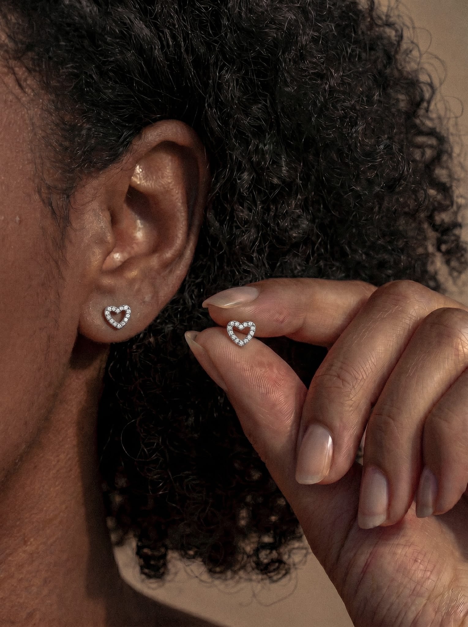 Primer plano de una persona que lleva el pendiente de botón «Love liso» con forma de corazón de Titanio-Noble Art, adornado con pequeños diamantes. Sostiene un pendiente a juego junto a la oreja, dejando entrever un piercing sobre un fondo neutro, lo que le da un toque elegante.