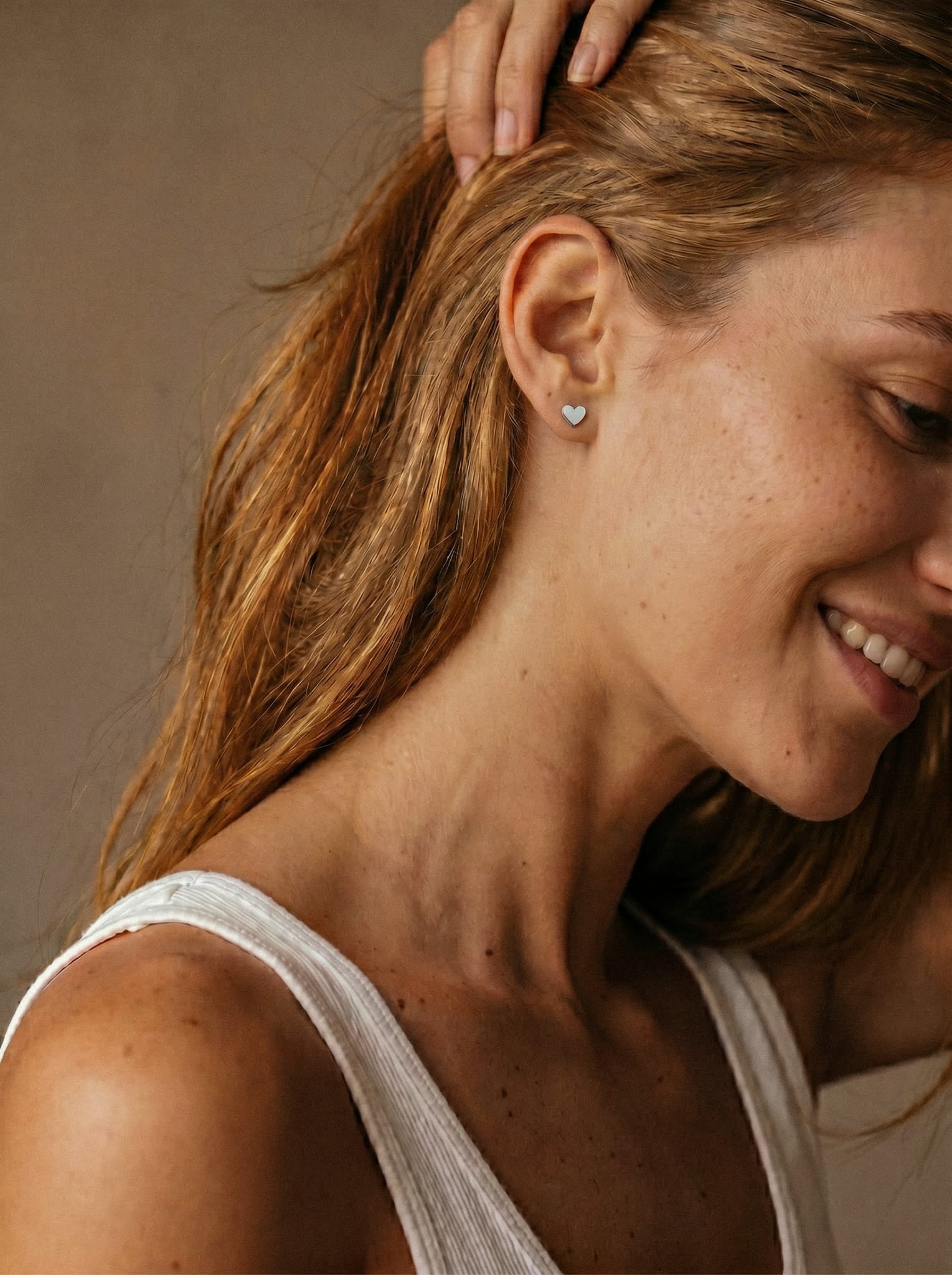 Una mujer de pelo castaño claro sonríe con una camiseta sin mangas blanca, luciendo el pendiente de botón en forma de corazón «LOVE LISO SILVER» de la colección «Noble Art» de Titanio. Se la ve de perfil sobre un fondo neutro.