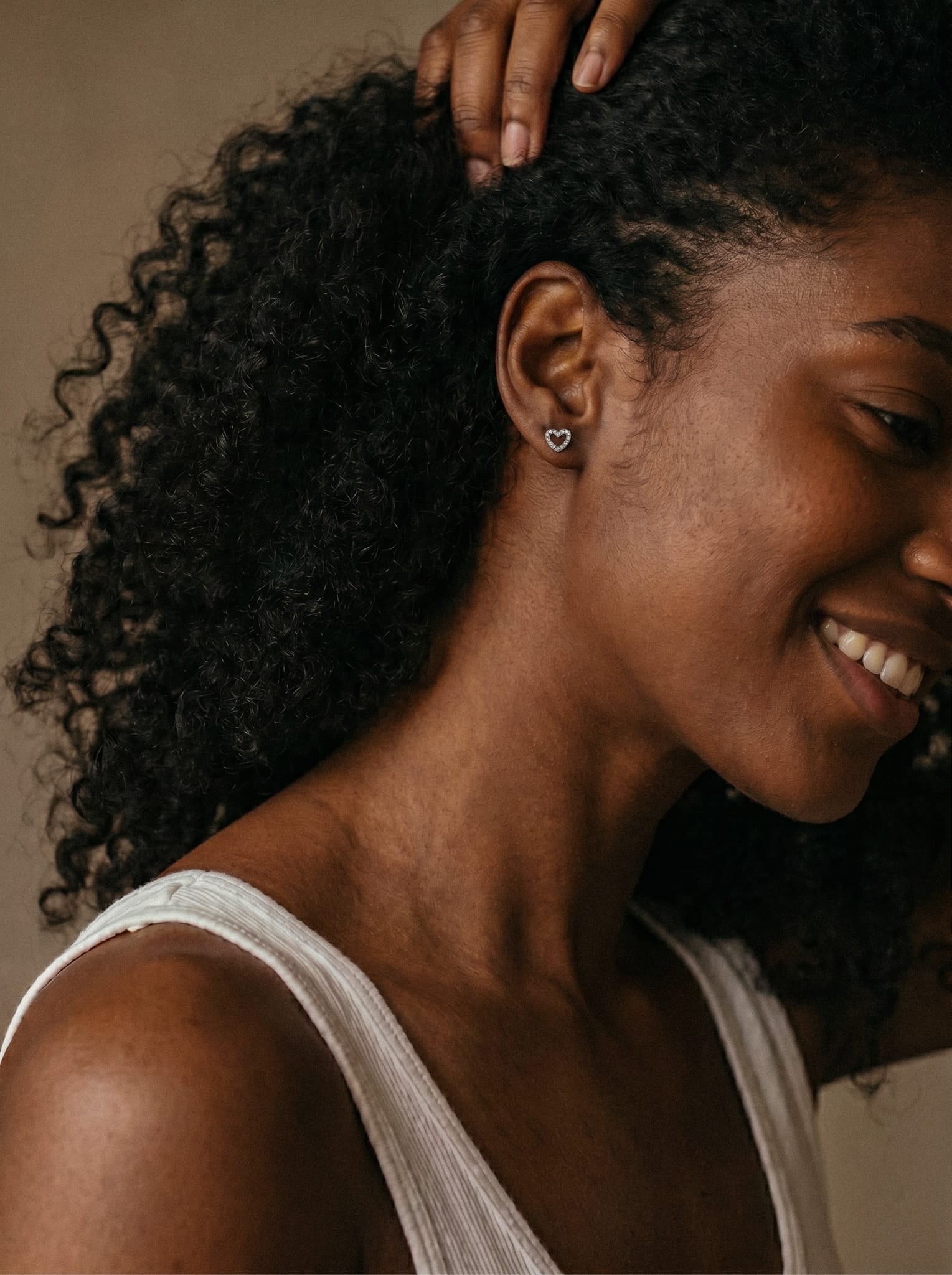 Una mujer de pelo rizado sonríe luciendo el pendiente «Love liso» de Titanio-Noble Art, una delicada pieza en forma de corazón que combina con un top blanco sin mangas y un elegante piercing.