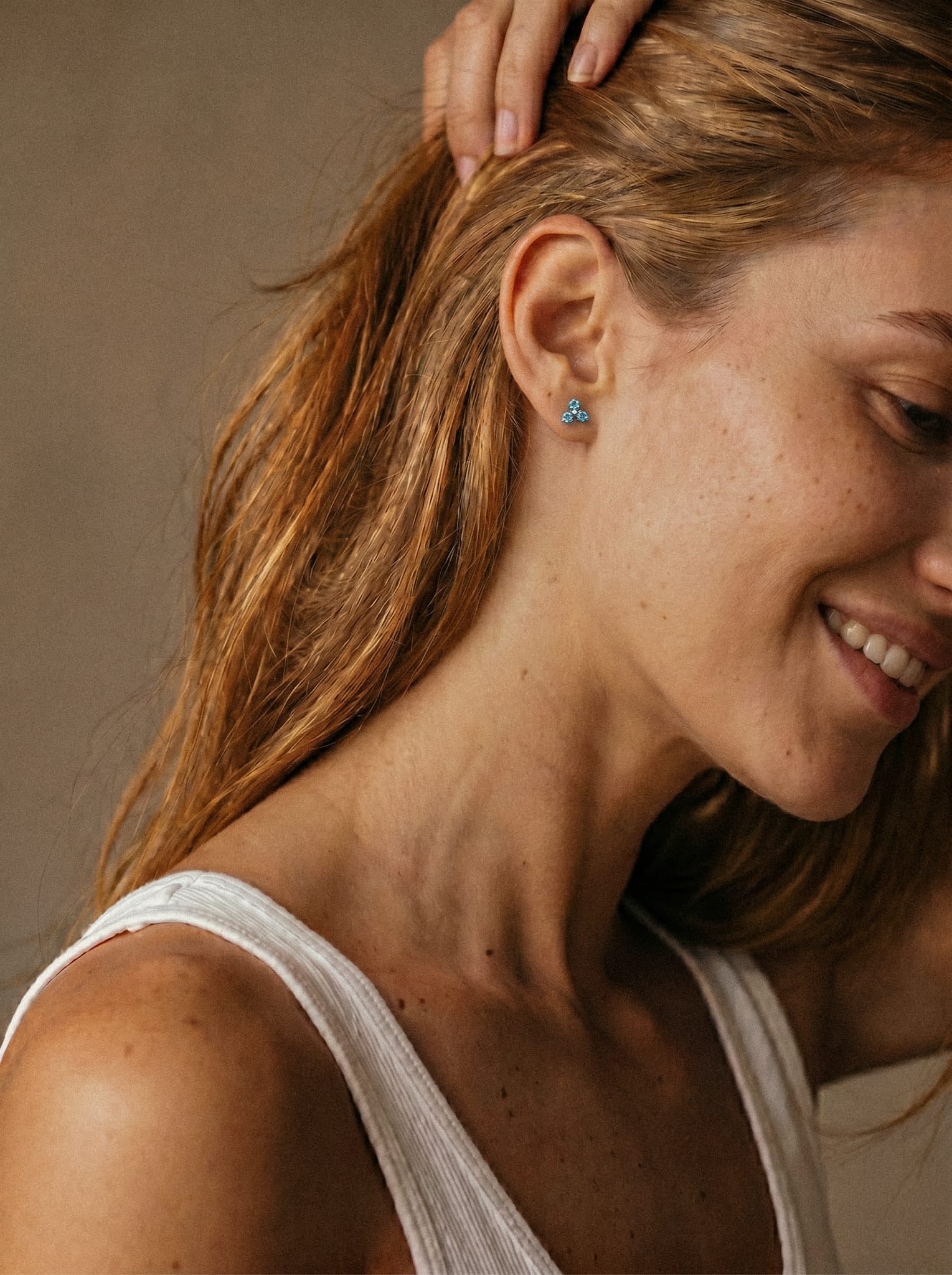 Una mujer de cabello castaño claro, con una camiseta blanca sin mangas, sonríe con dulzura; lleva un pendiente triangular azul pequeño «Trinity CZ» de Titanio-Noble Art que resalta su piercing sobre un fondo ligeramente difuminado.