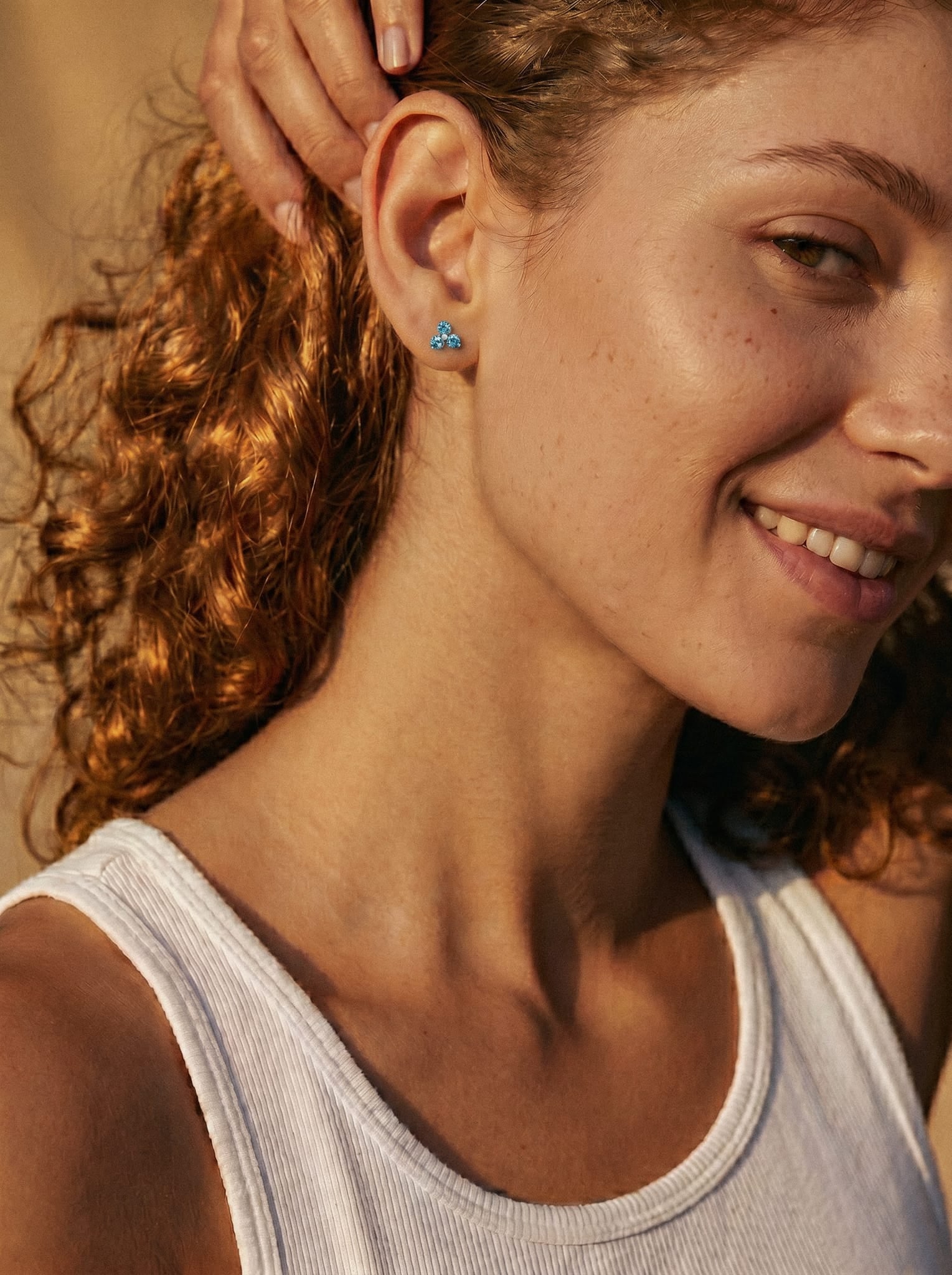 Una mujer de pelo rizado sonríe y se toca la oreja, mostrando el pendiente «Trinity CZ» de Titanio-Noble Art. Lleva una camiseta sin mangas blanca y aparece fotografiada bajo una luz cálida y natural.
