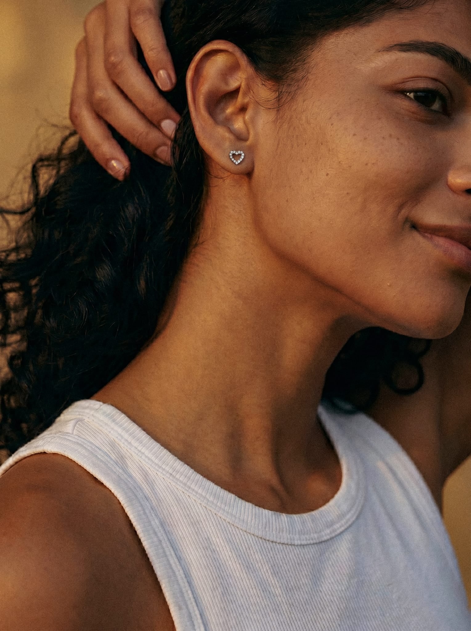 Una mujer con el pelo rizado lleva una camiseta blanca sin mangas y unos pendientes de botón con forma de corazón «Love liso» de Titanio-Noble Art; sonríe con dulzura y se toca el pelo sobre un fondo cálido inspirado en el estilo de tatuaje de Noble Art.