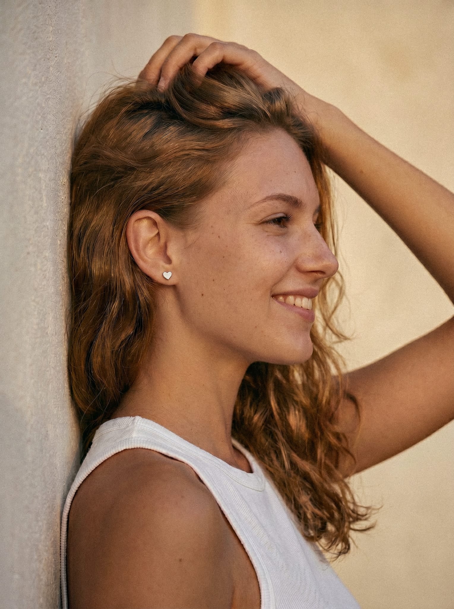 Una joven con el pelo castaño rojizo y ondulado, que lleva una camiseta blanca sin mangas y unos pendientes en forma de corazón, irradia confianza tras utilizar Titanio-Noble Art LOVE LISO SILVER. Se apoya contra una pared de tonos neutros, iluminada con luz tenue.