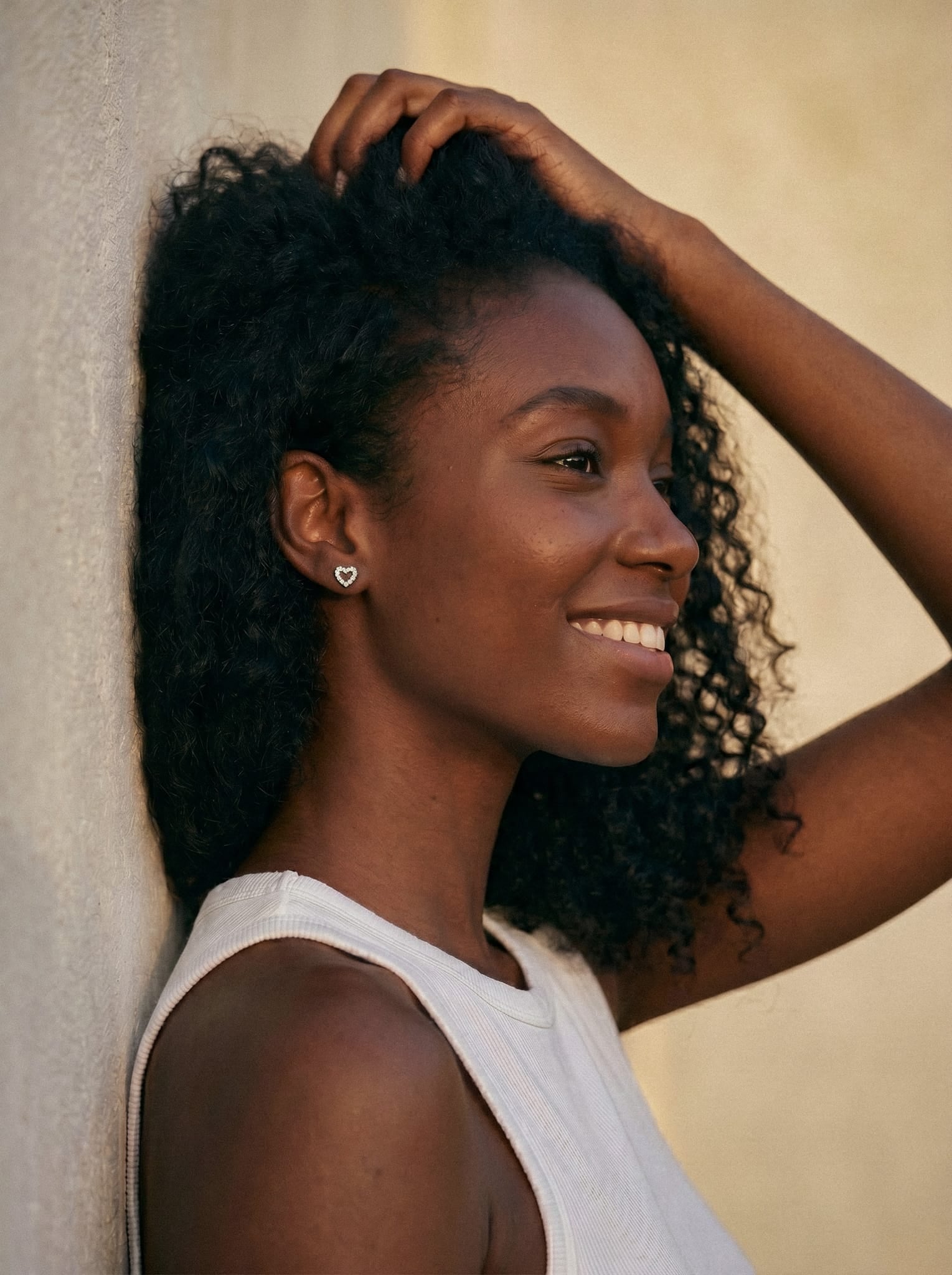 Una mujer con el pelo rizado sonríe, apoyada contra una pared clara y mirando hacia un lado. Lleva una camiseta blanca sin mangas, pendientes en forma de corazón y un delicado piercing. Descubre «Love liso» de Titanio-Noble Art.