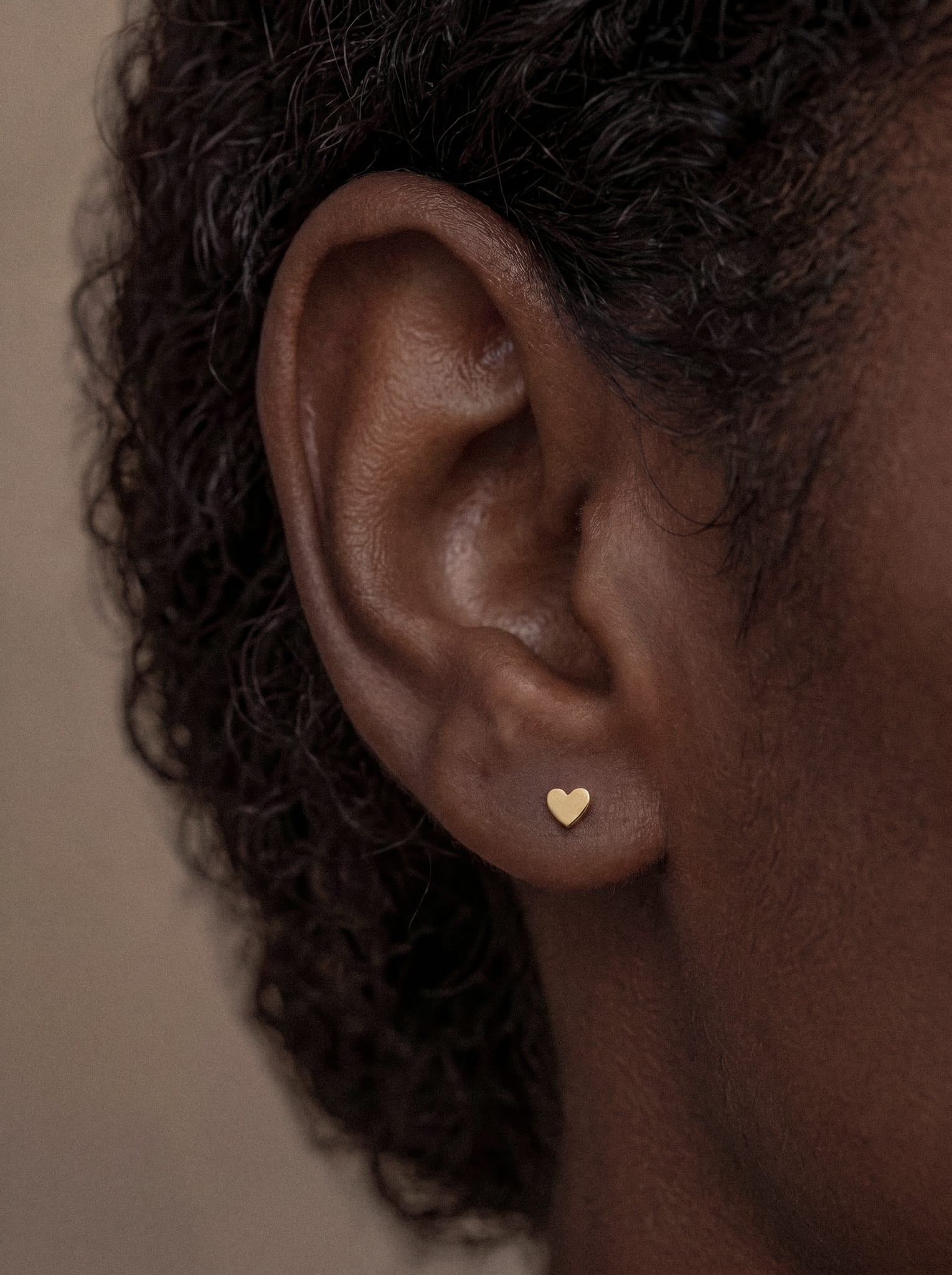 Close-up of a person’s ear wearing the Love liso small gold stud earring by Titanio- Noble Art. The image highlights the skin texture and curly hair near the ear against a neutral background, showcasing this piercing dorado as the regalo ideal for someone special.