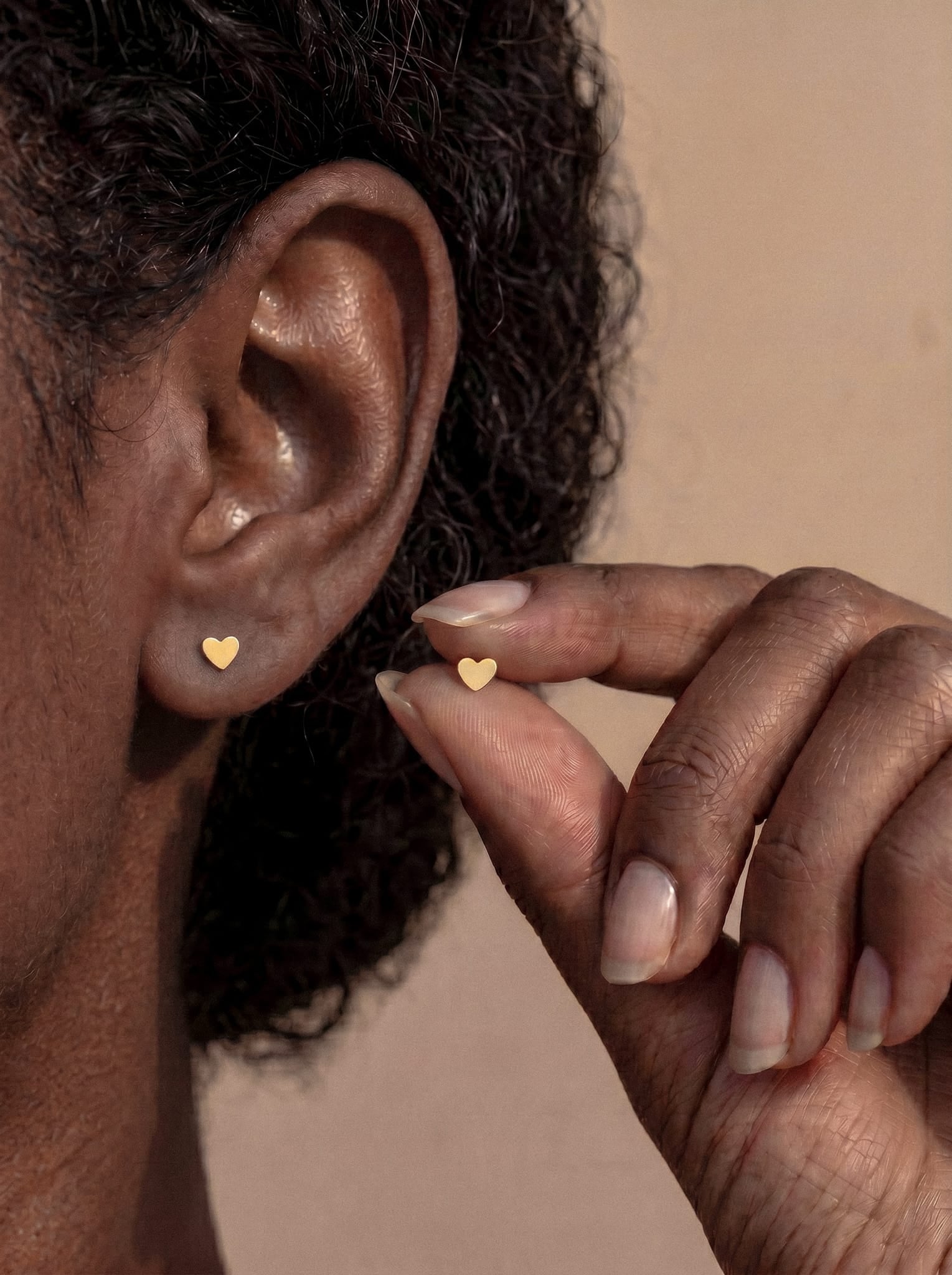 A close-up of a person’s ear adorned with the Love liso earring from Titanio- Noble Art—a petite dorado piercing shaped like a smooth heart. The person holds a matching earring near their ear, presenting it as el regalo ideal. The softly lit, neutral background highlights the elegance of the design.