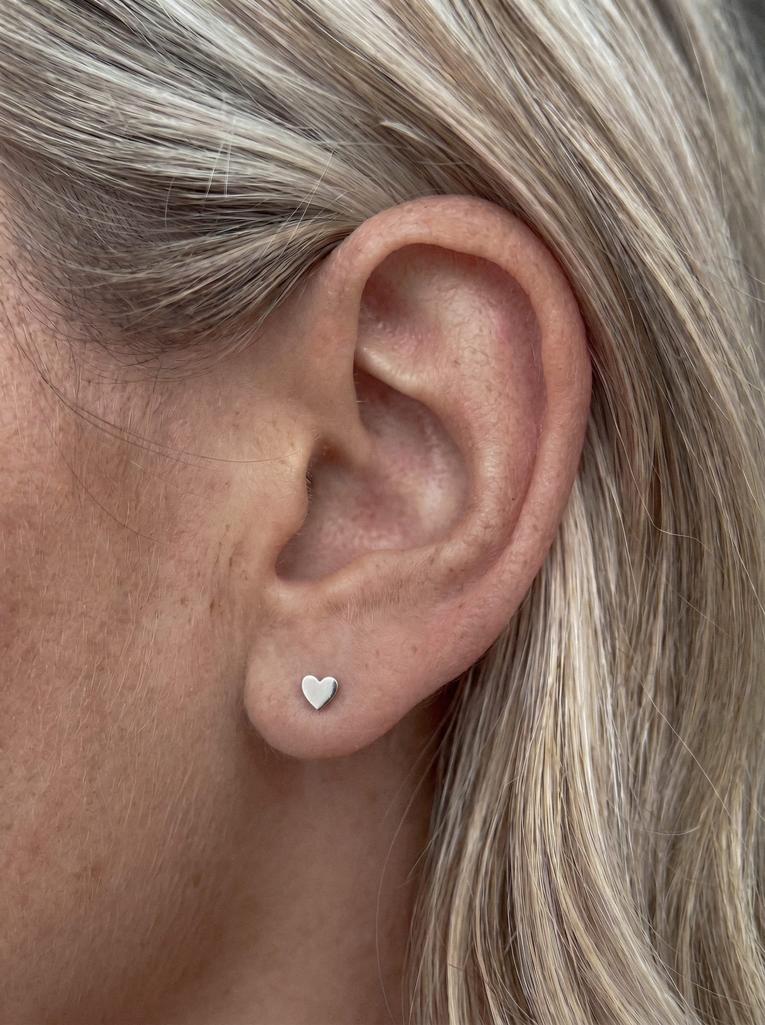 Close-up of a person’s ear with blonde hair, wearing the Love liso small silver heart-shaped stud earring by Titanio- Noble Art—the regalo ideal for someone special.