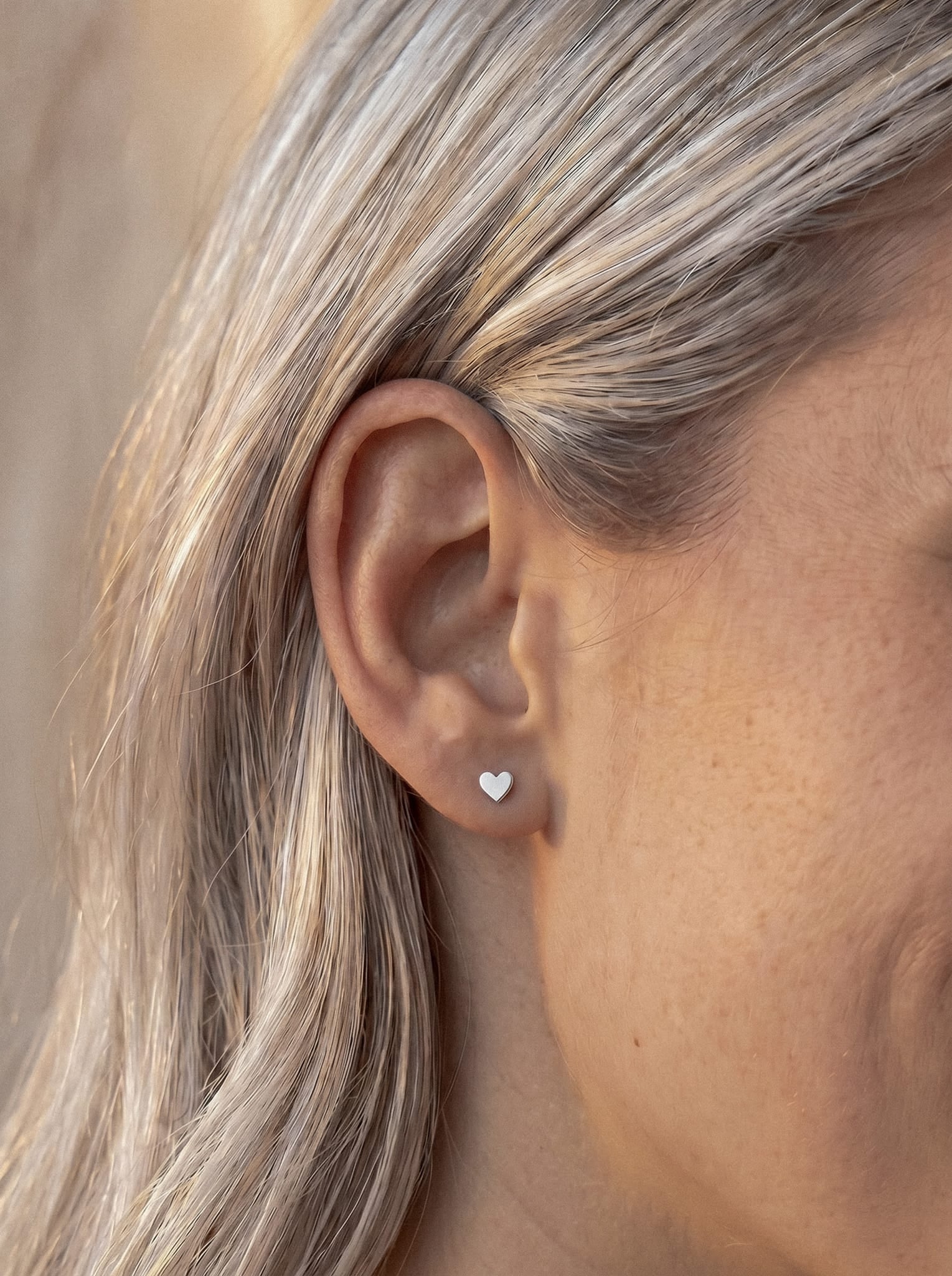 A close-up of a person’s ear with straight, light blonde hair, wearing the Love liso stud earring by Titanio- Noble Art—a small, silver corazón liso that adds an elegant touch, making it a regalo ideal.