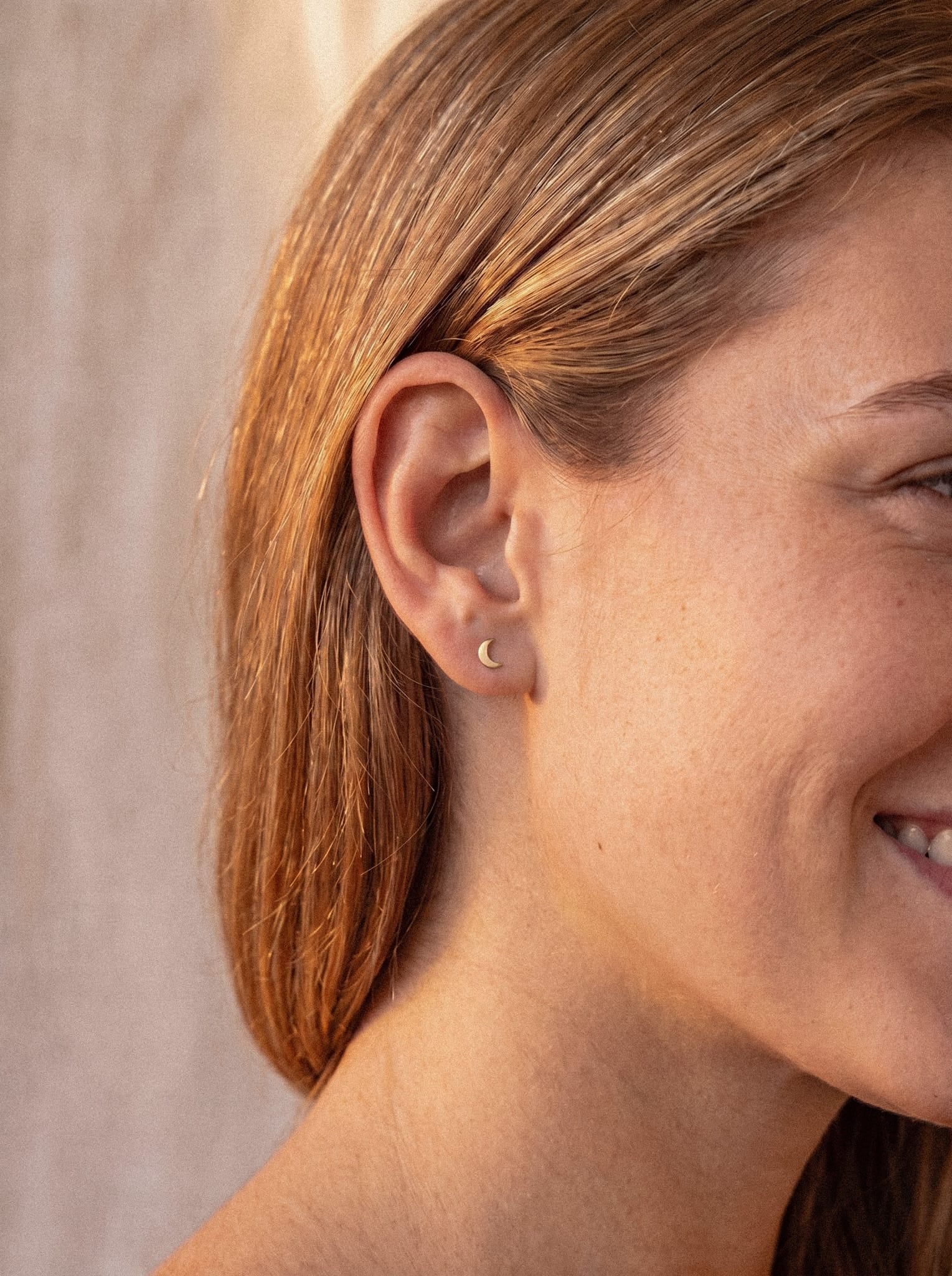 Primer plano de una mujer sonriente, con el pelo liso y castaño claro, que luce el pendiente «Luna liso» de Titanio-Noble Art; su delicada forma de media luna dorada es un sutil guiño a la geometría lunar. El fondo neutro, iluminado con luz suave, resalta la elegancia discreta de la pieza.