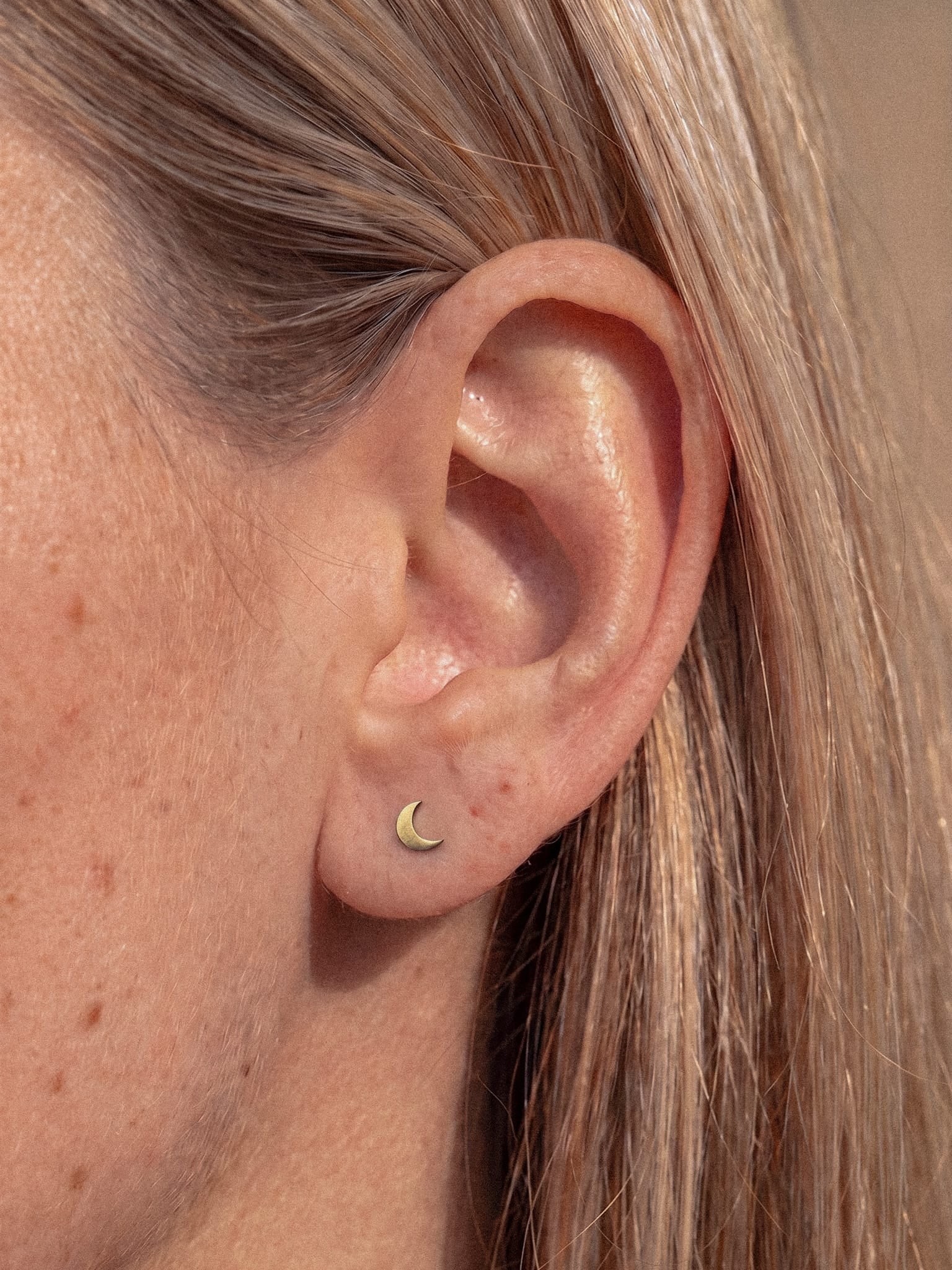 Primer plano de la oreja de una persona con el pelo rubio y liso, que lleva el pendiente de botón «Luna liso», pequeño y dorado con forma de media luna, de la marca Titanio-Noble Art, inspirado en la geometría lunar.