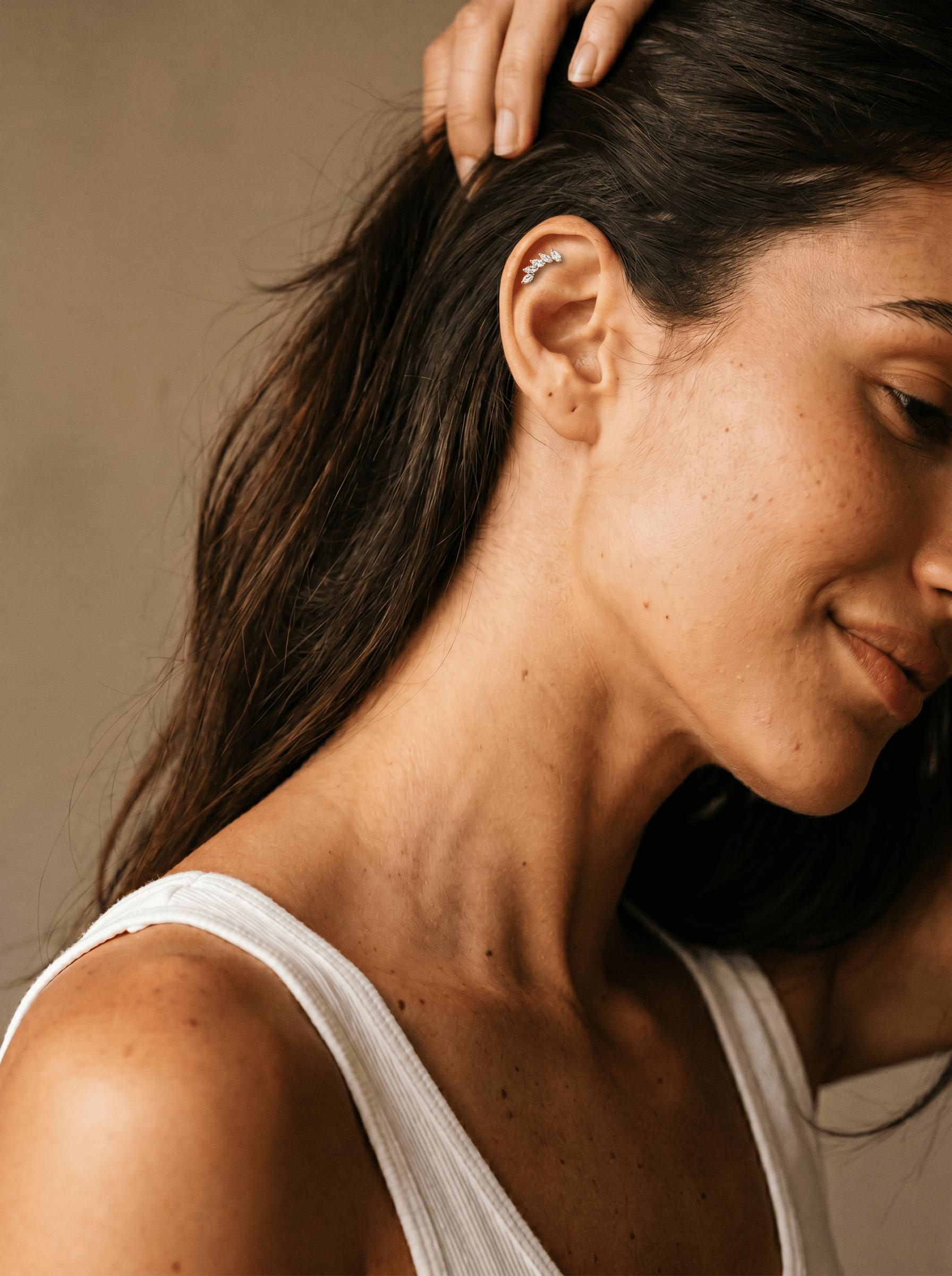 Una mujer de pelo largo y castaño, con una leve sonrisa y vestida con una camiseta sin mangas blanca, se toca la cabeza para mostrar varios pendientes de aro «Gia Dorado» de Titanio-Noble Art sobre un fondo neutro y elegante.