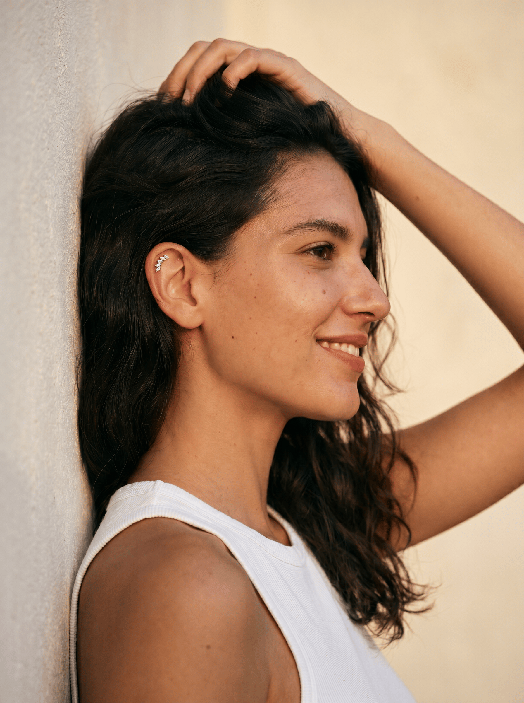 Una mujer con el pelo largo, castaño y ondulado, y con piercings en las orejas, sonríe con seguridad mientras se apoya contra una pared clara, con la mano en la cabeza. Luce el modelo «Gia dorado» de Titanio-Noble Art, quizá pensando en la eliminación láser de tatuajes.