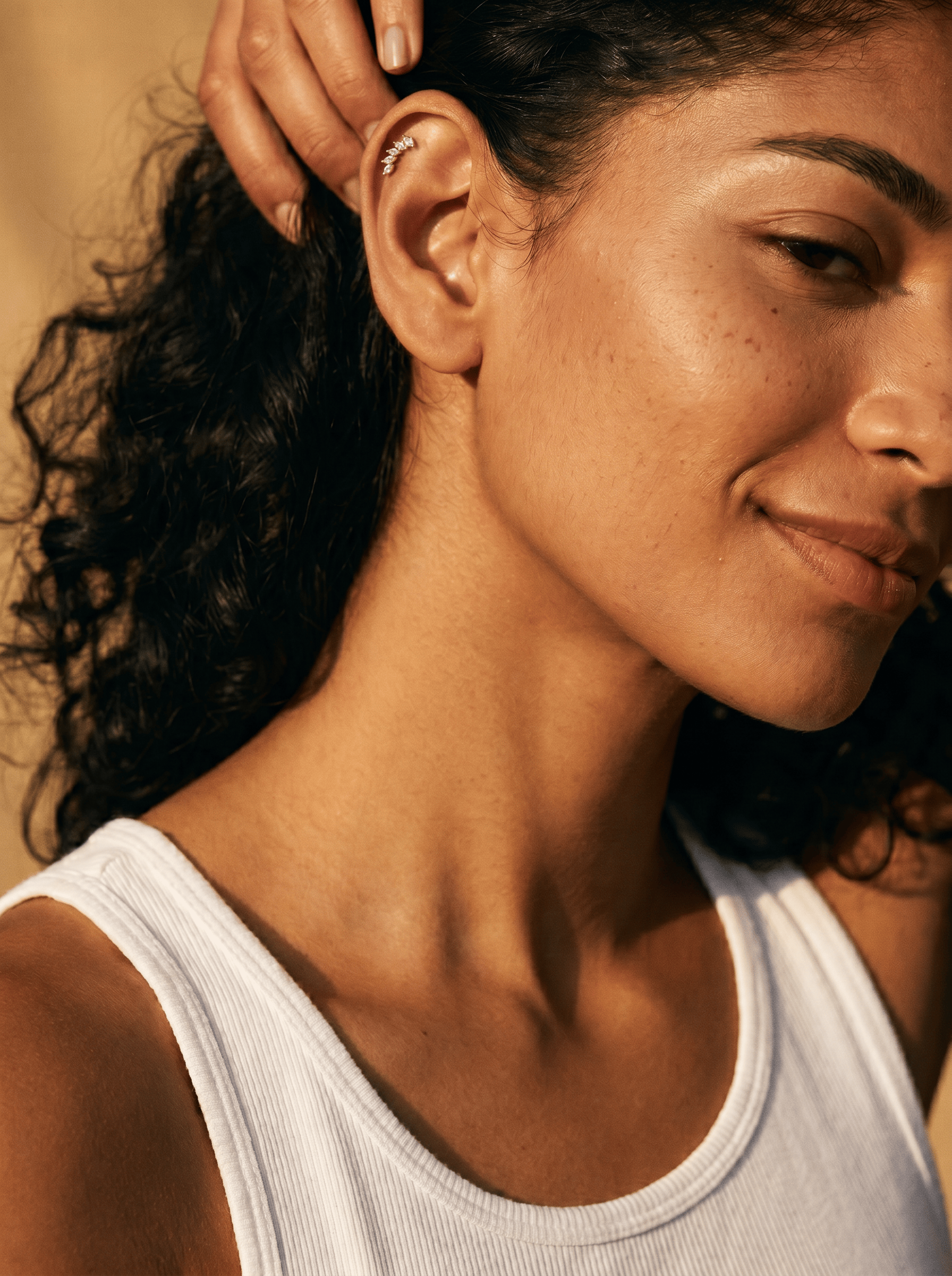 Una mujer de pelo rizado sonríe con dulzura, vestida con una camiseta sin mangas blanca. Levanta la mano para mostrar el elegante pendiente tipo «ear cuff» Gia dorado de Titanio-Noble Art, y la luz resalta su piel clara y su belleza natural.