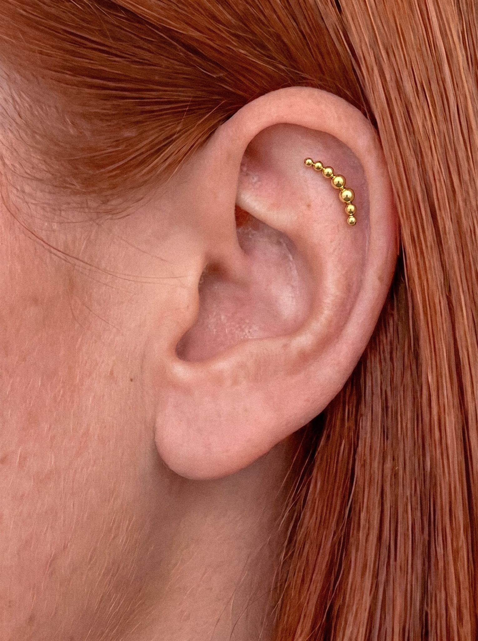 Un primer plano de una oreja con vello rojizo, en el que se aprecia la joya de cartílago «Moon cluster» de Titanio-Noble Art, con cuentas de oro, colocada a lo largo de la curva exterior de la oreja.