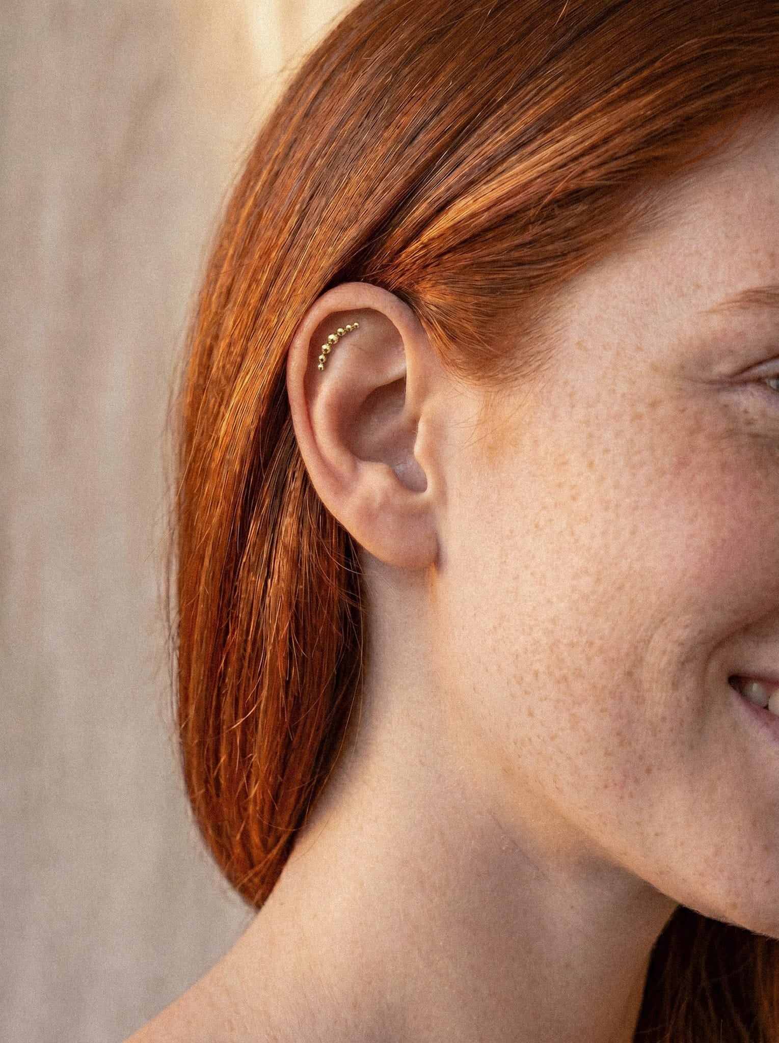 Un primer plano de una mujer pelirroja y con pecas, sonriendo. Lleva puestos los pendientes «Moon cluster» de Titanio-Noble Art a lo largo del borde de la oreja, lo que pone de manifiesto un estilo minimalista en los piercings de joyería.