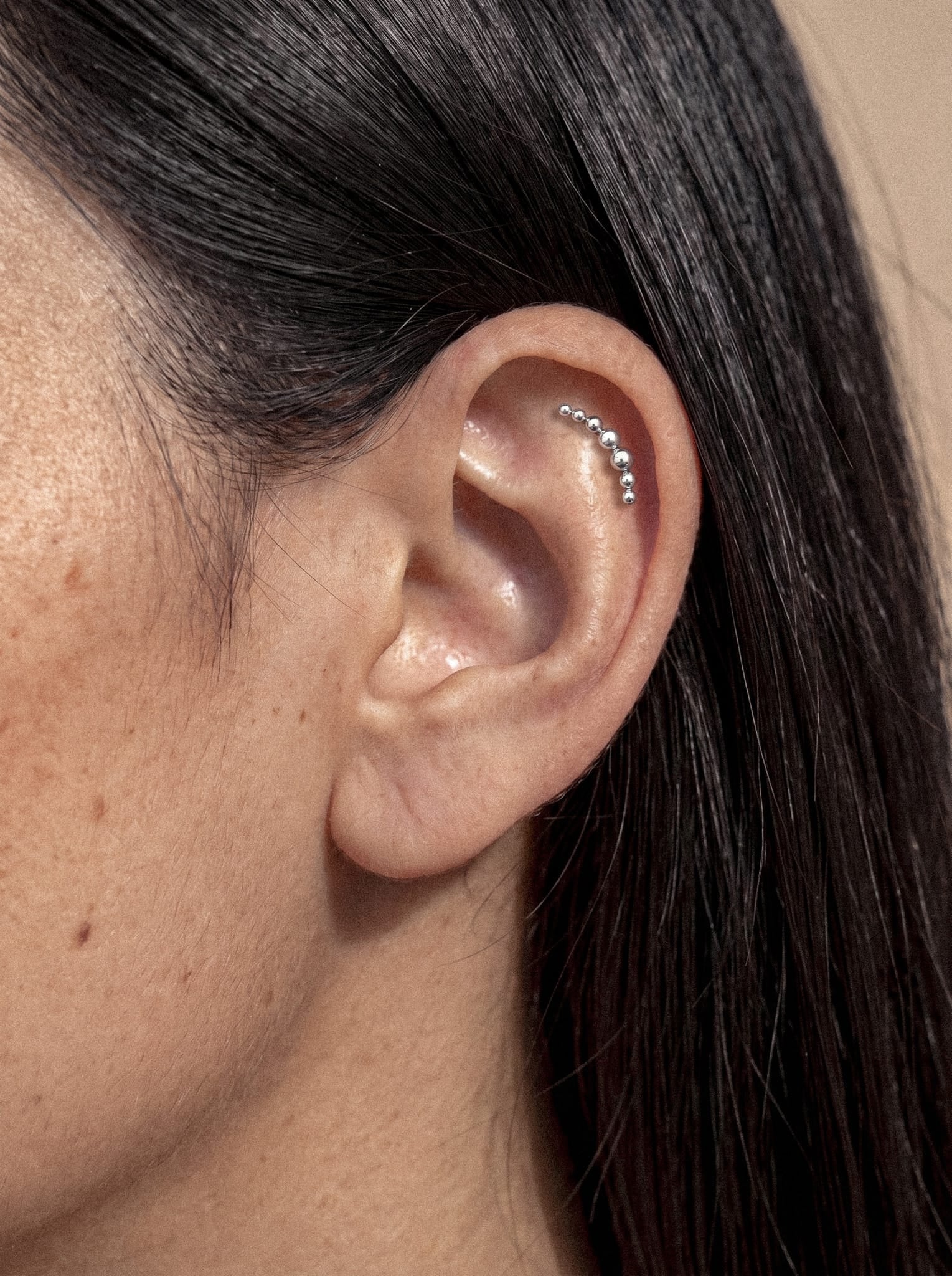 Un primer plano de la oreja de una persona, adornada en el cartílago con el «Moon Cluster» de Titanio-Noble Art —una fila curvada de pequeños pendientes de plata—, que pone de manifiesto el minimalismo en los piercings. La persona tiene el pelo liso y de color castaño oscuro.