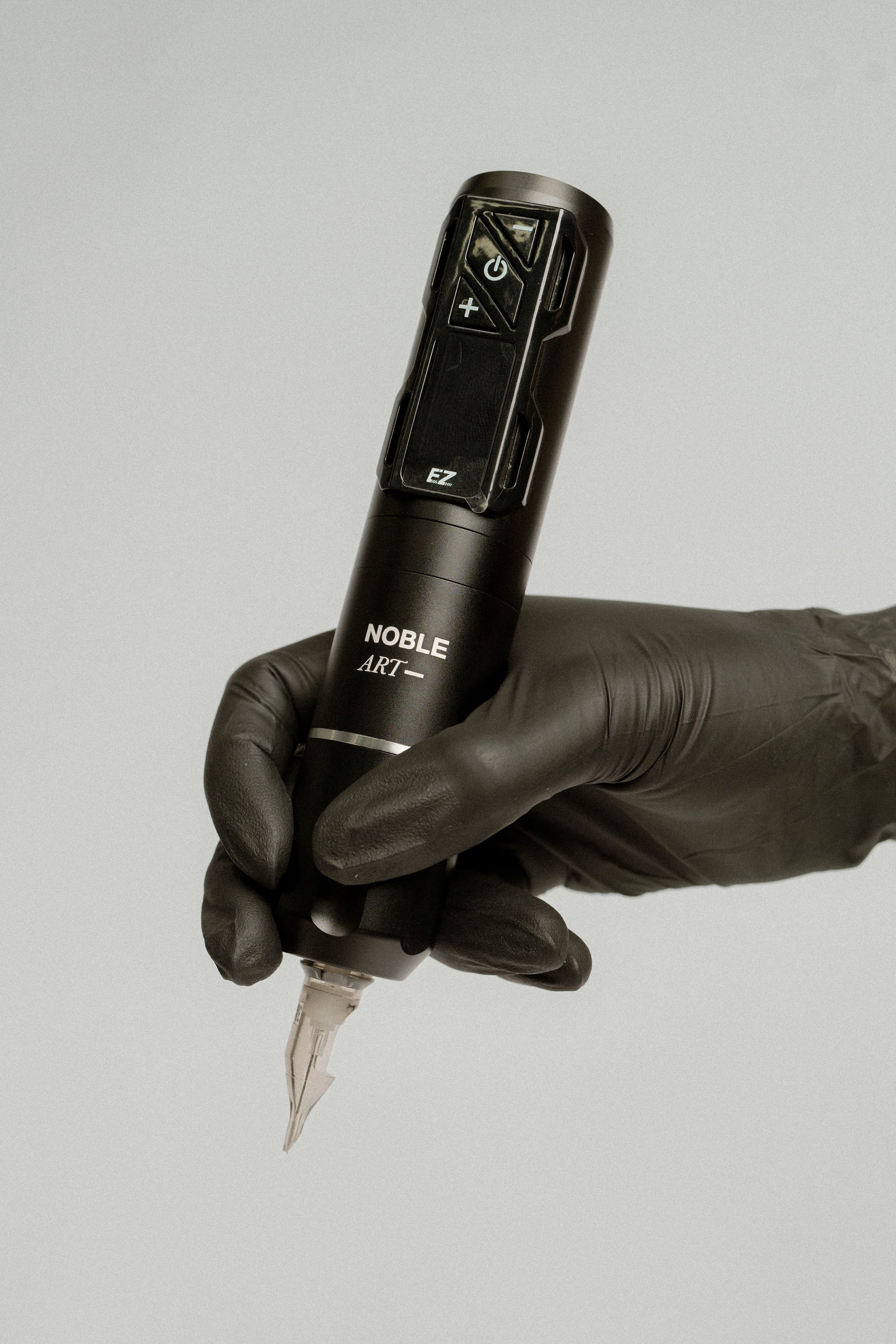 Una mano enguantada sostiene la «Máquina de tatuar rotativa inalámbrica EZ 2PRO Wireless» de Noble Art, ideal para tatuajes y piercings, sobre un fondo liso de color gris claro.