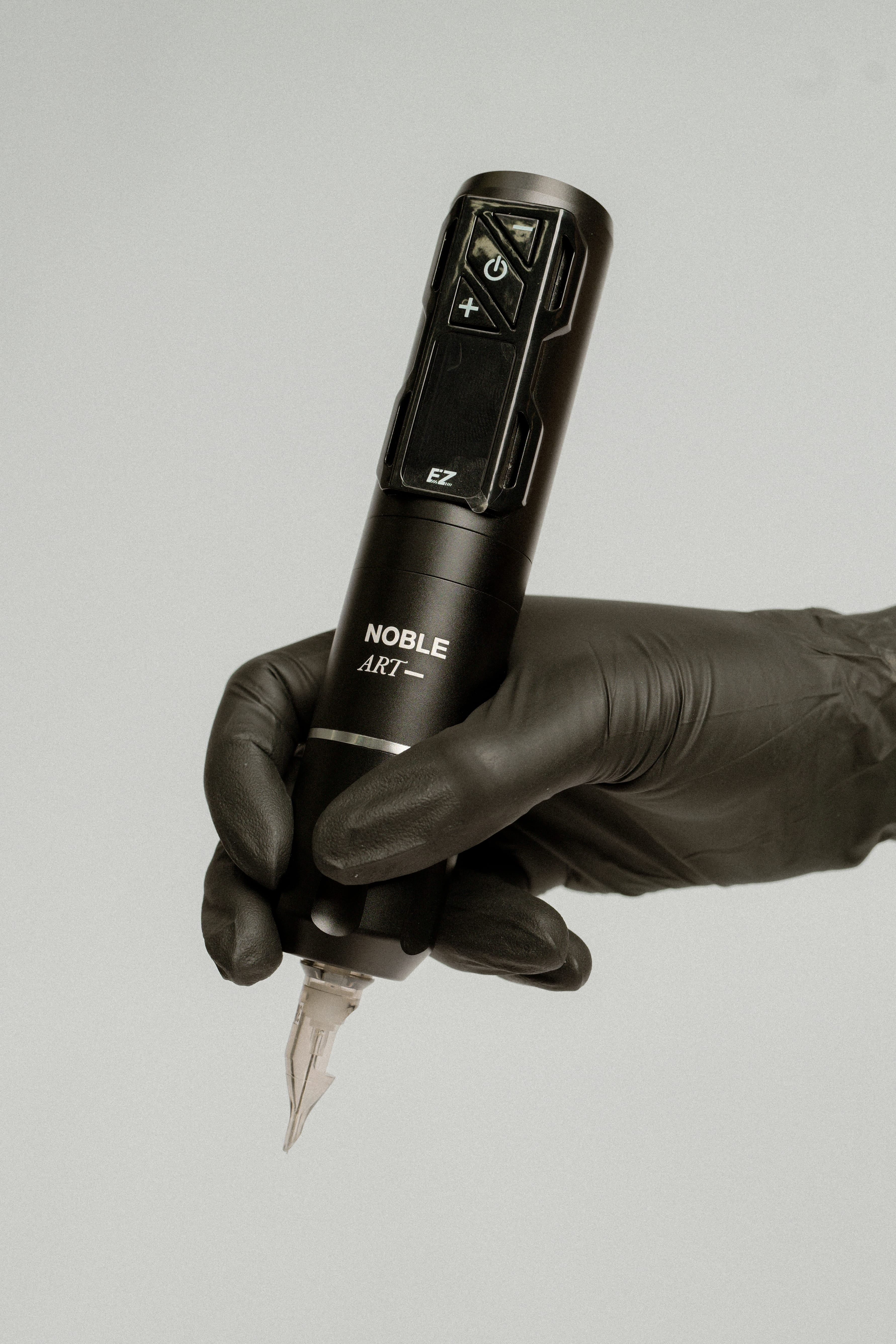 Una mano enguantada sostiene la «Máquina de tatuar rotativa inalámbrica EZ 2PRO Wireless» de Noble Art, ideal para tatuajes y piercings, sobre un fondo liso de color gris claro.