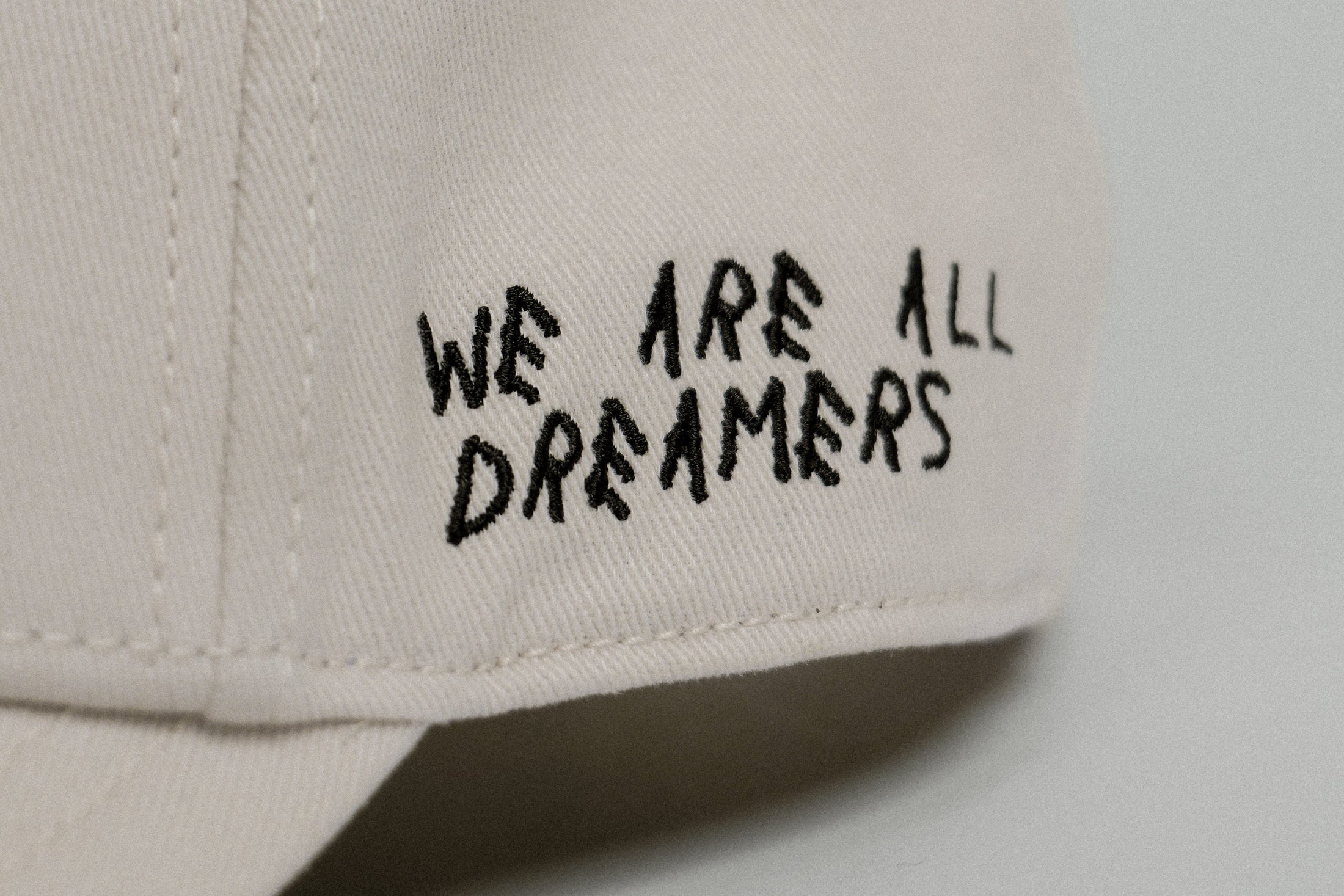 Close-up of the GORRA NOBLE ART: a beige fabric cap with "WE ARE ALL DREAMERS" embroidered in black, uneven letters on the side—inviting self-expression and personal style transformation.