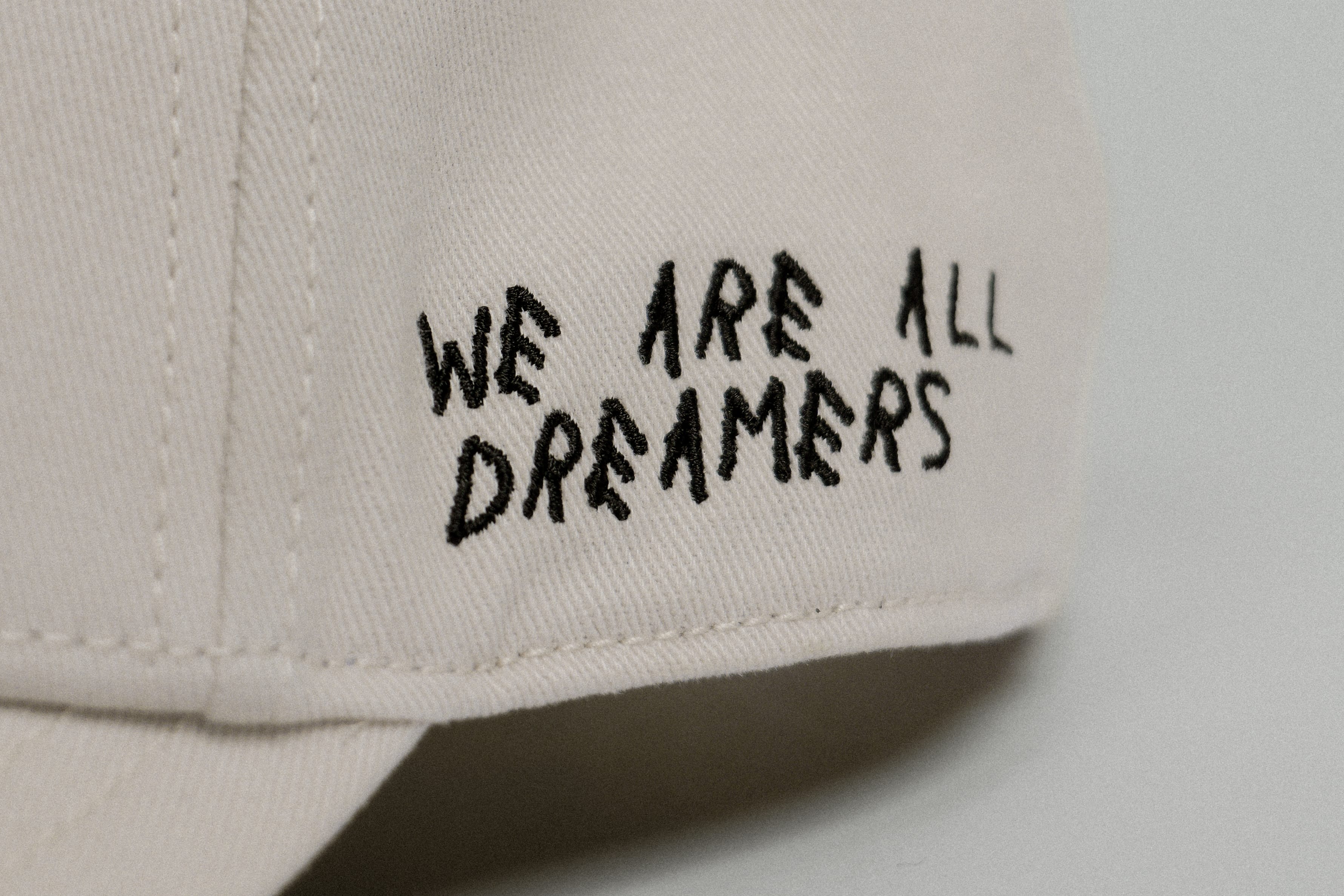 Close-up of the GORRA NOBLE ART: a beige fabric cap with "WE ARE ALL DREAMERS" embroidered in black, uneven letters on the side—inviting self-expression and personal style transformation.