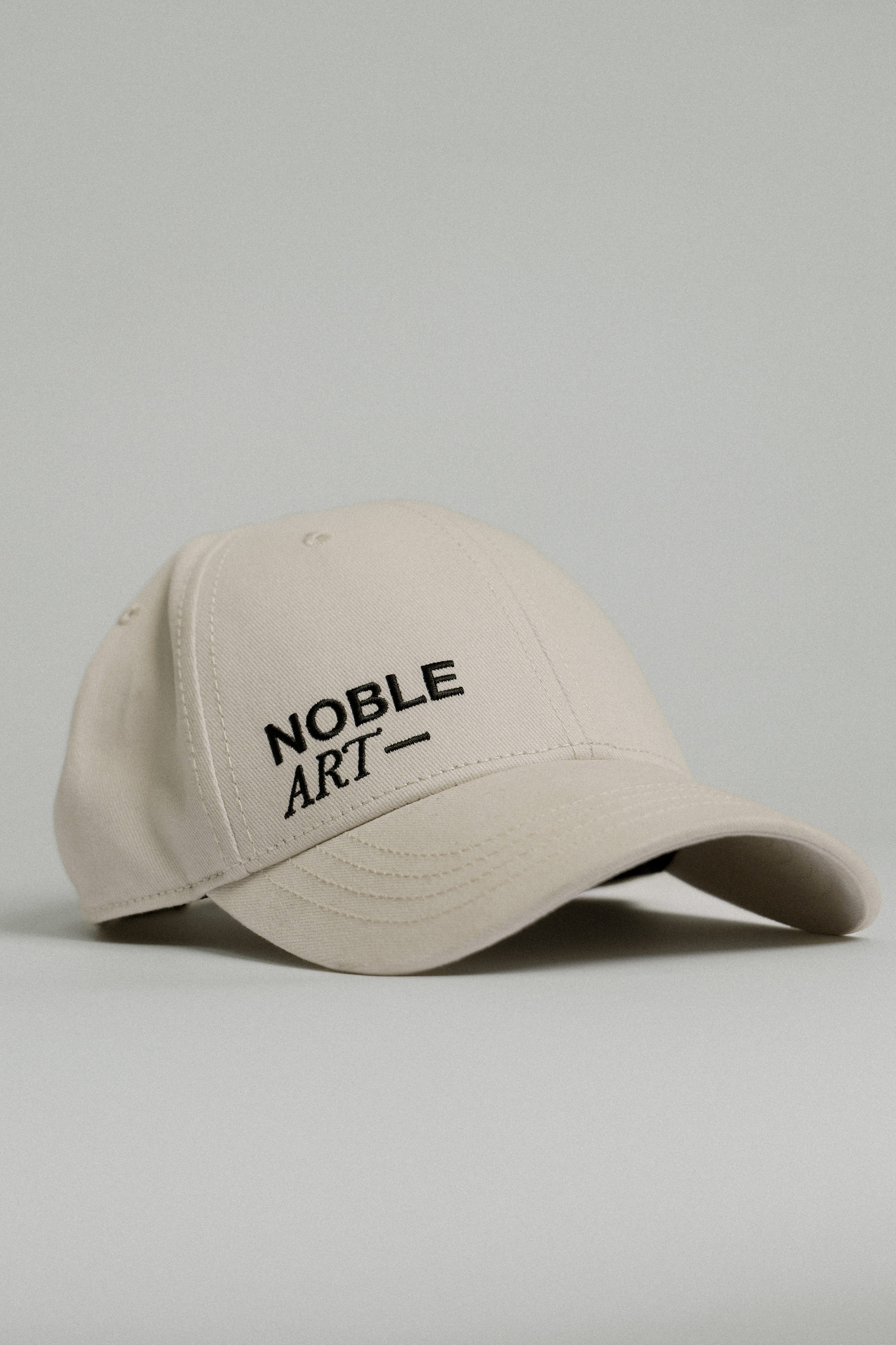 The GORRA NOBLE ART is a beige baseball cap featuring "NOBLE ART" embroidered in black on the front, inspired by Noble Art Miami and shown against a plain light gray background.