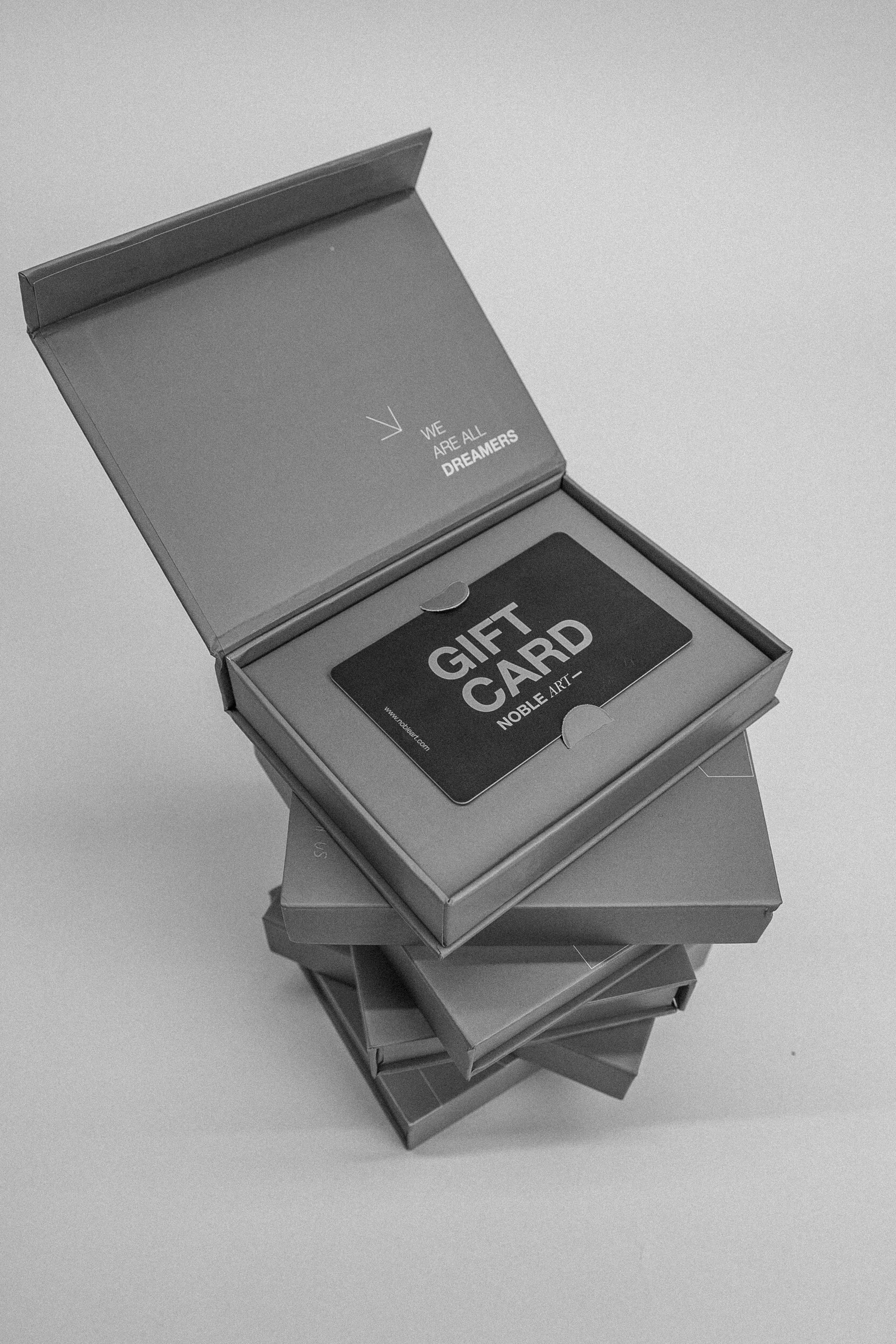 Se muestra una tarjeta regalo de 50 de A NOBLE ART BEAUTY SOCIEDAD LIMITADA en una caja abierta, con más cajas apiladas debajo. La imagen en blanco y negro muestra una tapa en la que se lee: «Todos somos soñadores», lo que refleja el noble arte de hacer regalos.
