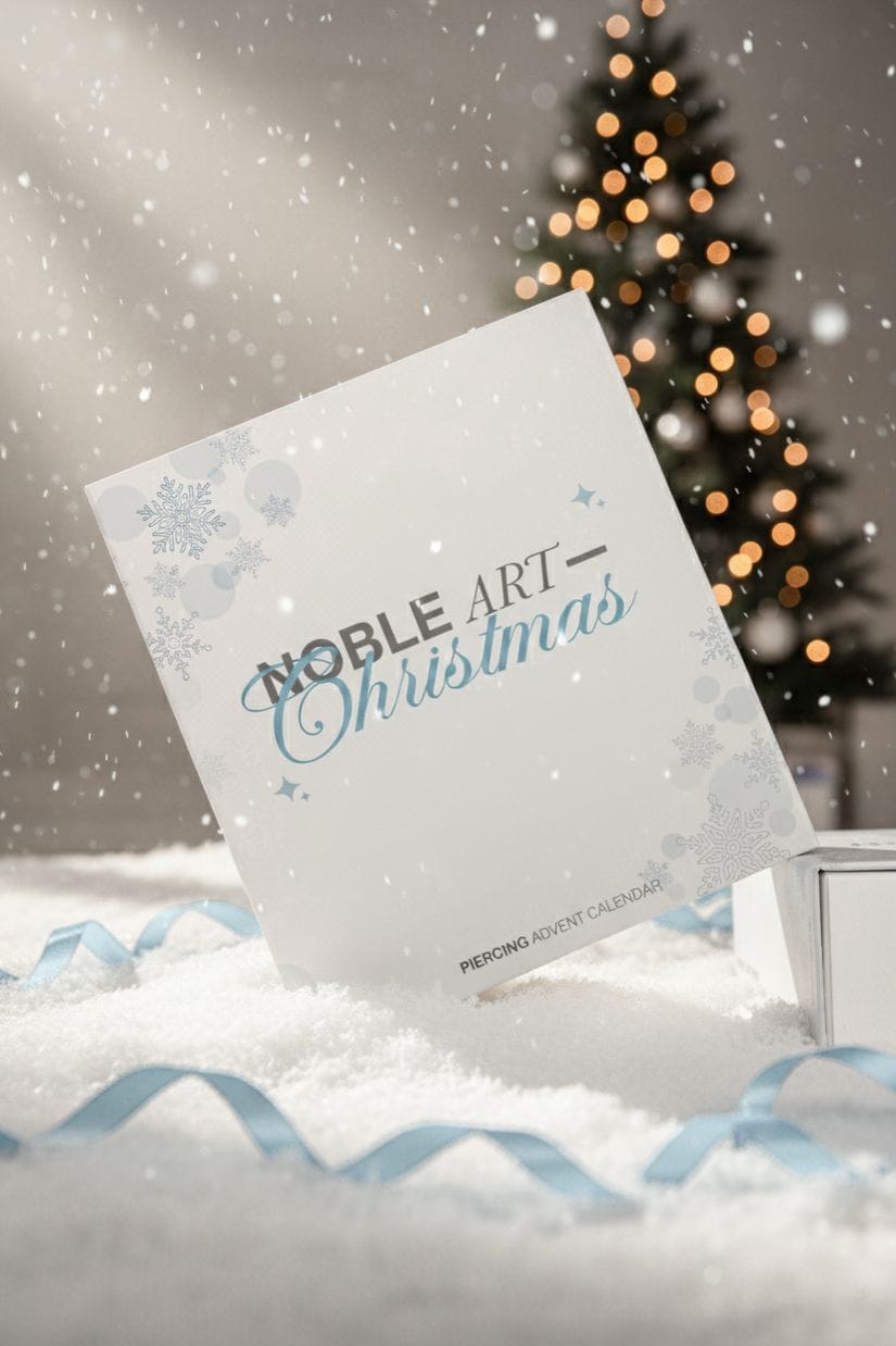 Una tarjeta blanca en la que se lee «Calendario de Adviento Noble Art», de NOBLE ART BEAUTY SOCIEDAD LIMITADA, se alza sobre la nieve, con una cinta azul junto a ella. Un árbol de Navidad iluminado brilla suavemente en el fondo difuminado, mientras la nieve cae alrededor de este regalo que llama la atención.