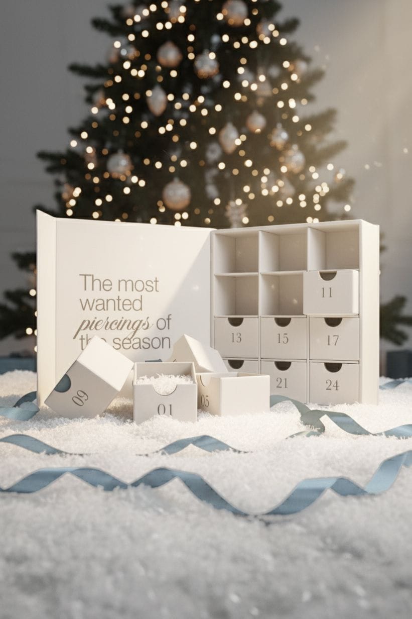El calendario de Adviento Noble Art de NOBLE ART BEAUTY SOCIEDAD LIMITADA, con cajitas numeradas, aparece abierto sobre la nieve, frente a un árbol de Navidad decorado. Una de las cajitas deja al descubierto papel de seda en su interior y en la caja se lee: «Los piercings más buscados de la temporada».