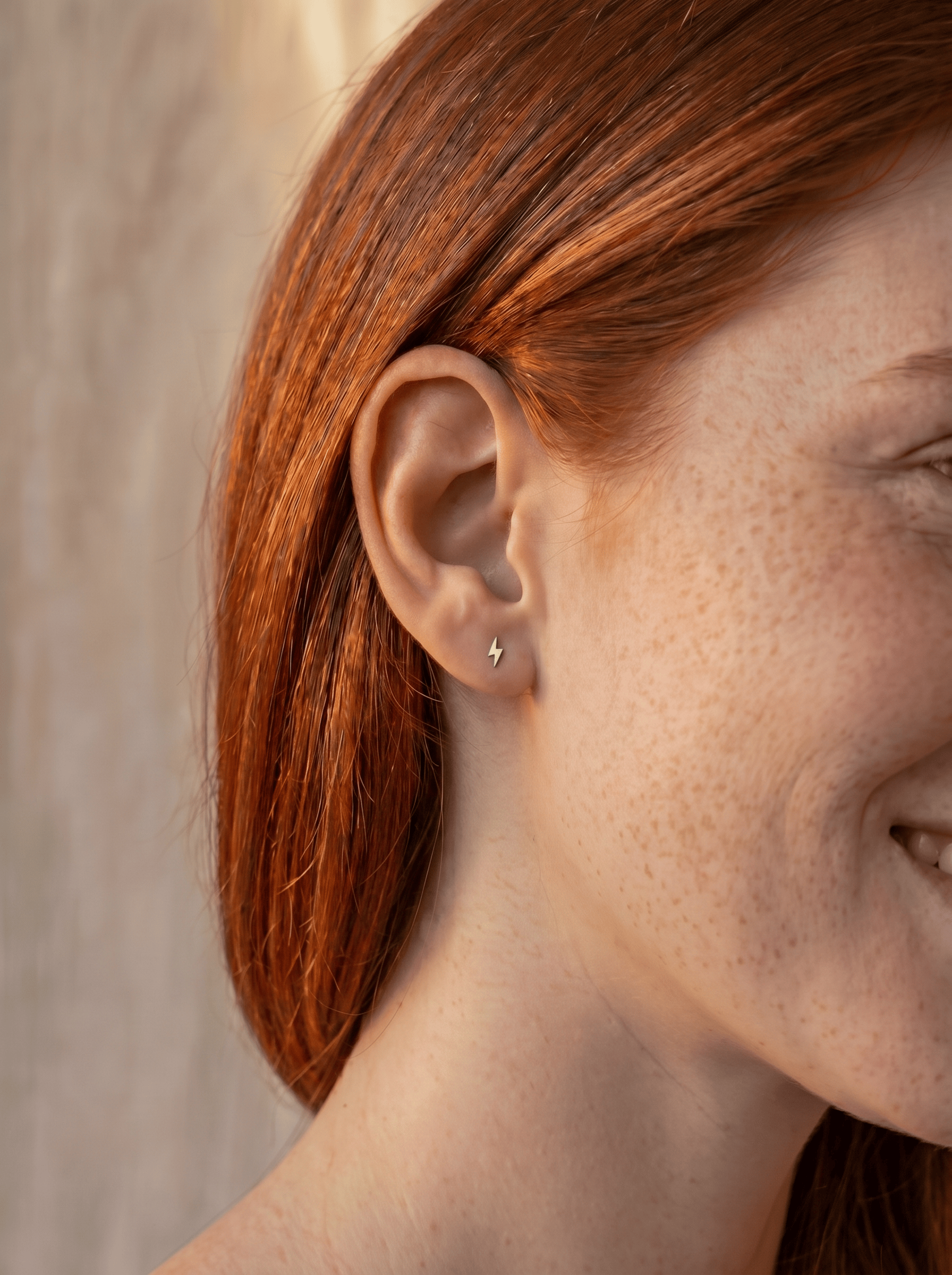 Primer plano de una persona sonriente, con el pelo largo y rojo y pecas, que lleva en la oreja izquierda el «Rayo» de Titanio-Noble Art —un pequeño pendiente plateado con forma de rayo—. El fondo, ligeramente desenfocado y en tonos neutros, acentúa la energía pura y el estilo minimalista de la escena.