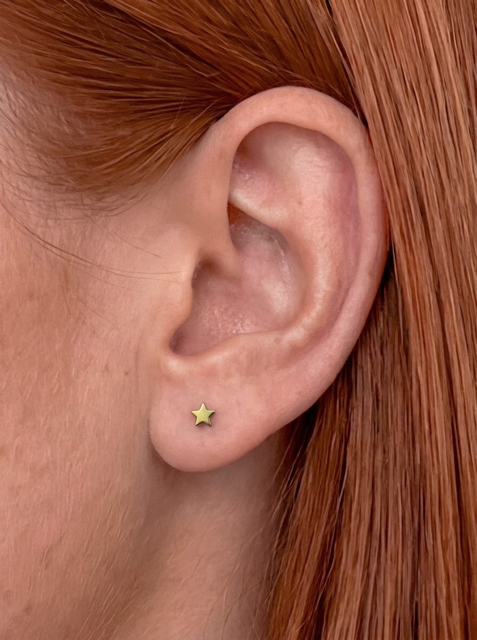 Primer plano de la oreja de una persona con el pelo liso y rojizo. La oreja luce el pendiente «Star liso» de Titanio-Noble Art en el lóbulo, lo que aporta un delicado toque de joyería a su look.