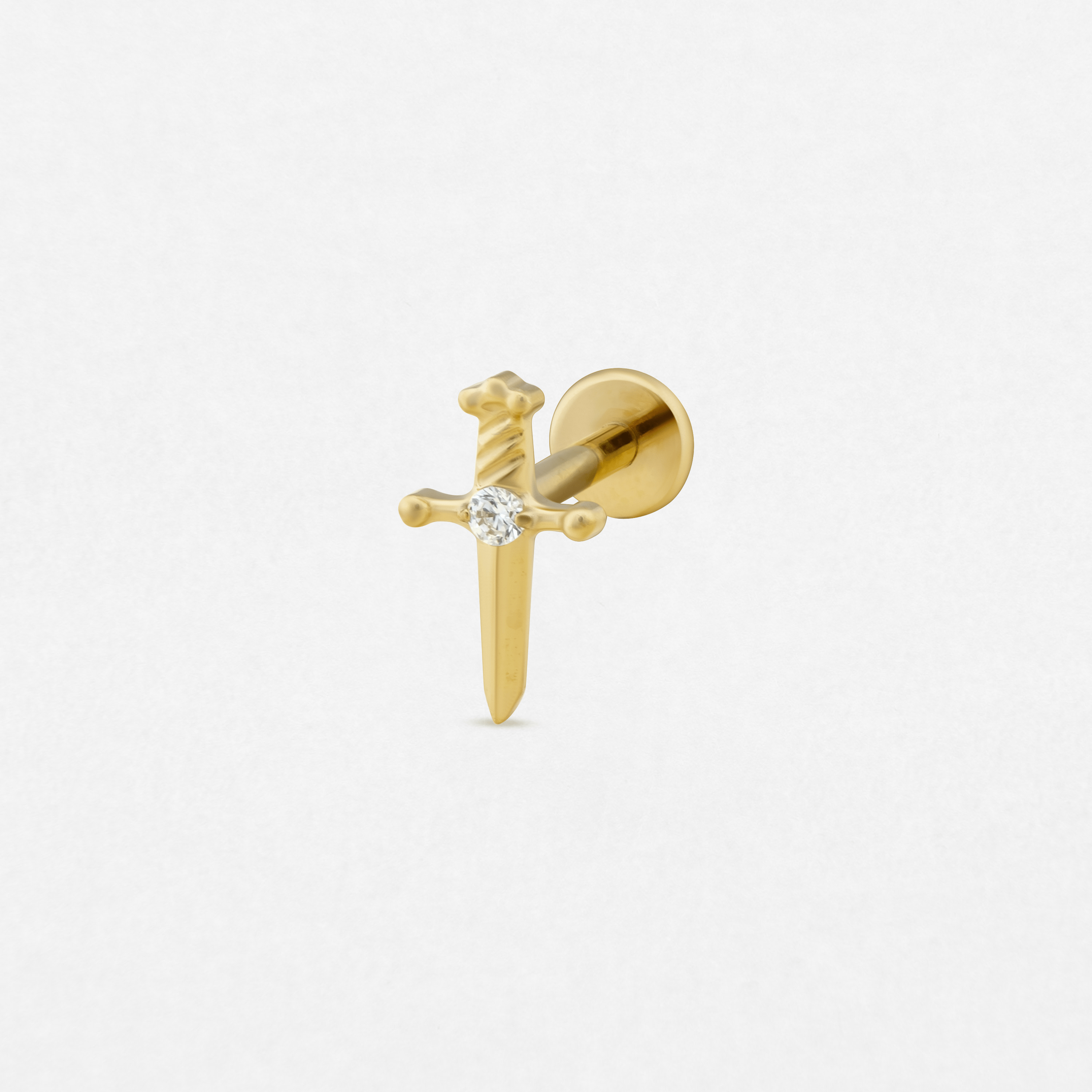 El pendiente de oro «Daga» de Titanio-Noble Art tiene forma de espada con una gema redonda y transparente en la guarda, y se presenta sobre un fondo blanco liso.