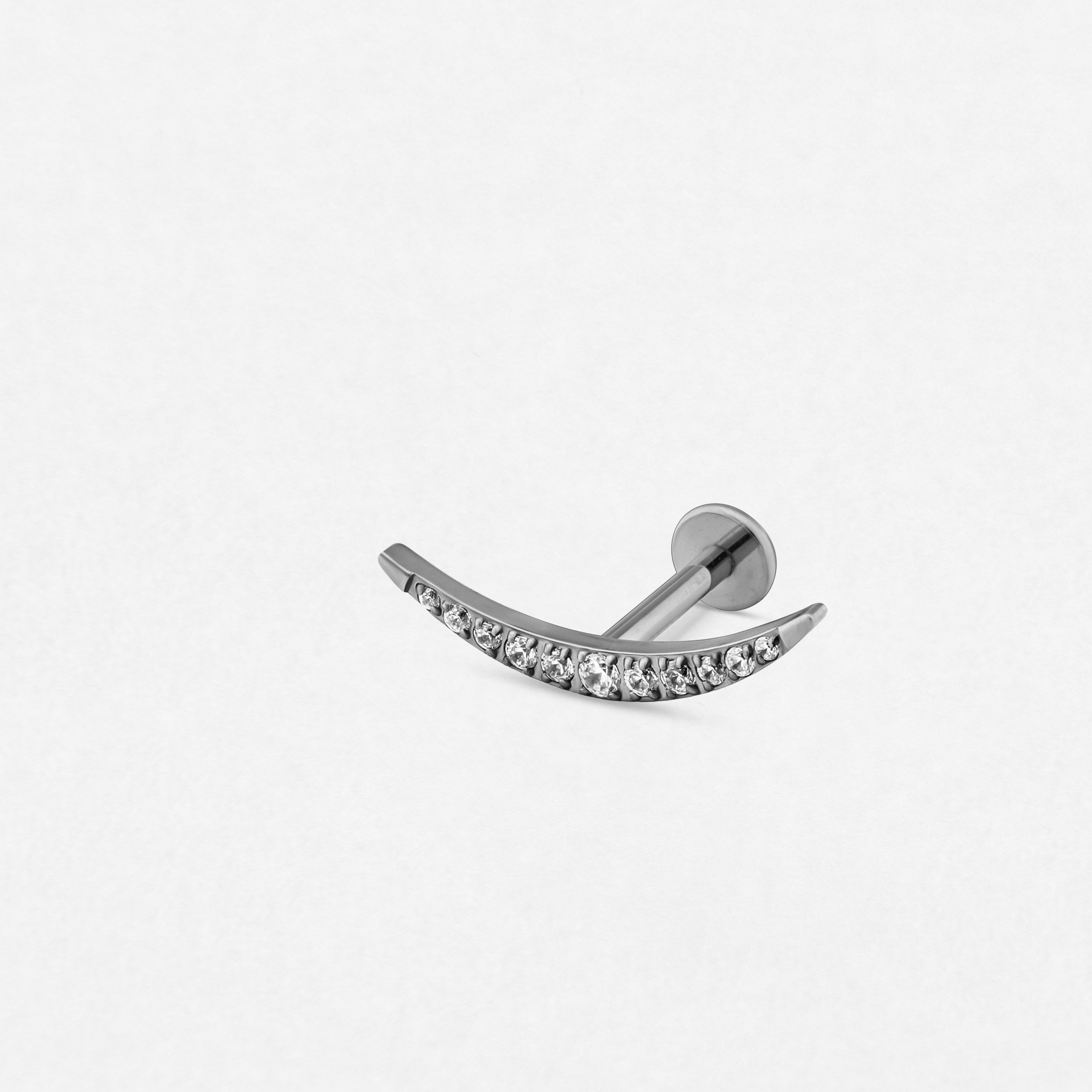 El modelo «Atenea Silver» de Titanio-Noble Art es un pendiente tipo barbell de plata con forma curvada, adornado con piedras preciosas transparentes y presentado sobre un fondo blanco; ideal para los amantes de las joyas corporales elegantes o para quienes estén pensando en someterse a una futura eliminación con láser.