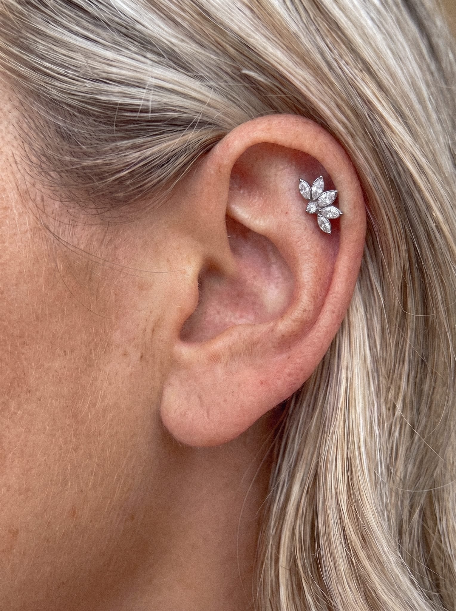 Primer plano de la oreja de una persona con el pelo rubio, adornada con el pendiente de cartílago «Ava» de Titanio-Noble Art: una pieza de plata decorada con múltiples cristales en forma de pétalo dispuestos en una joya con forma de flor.