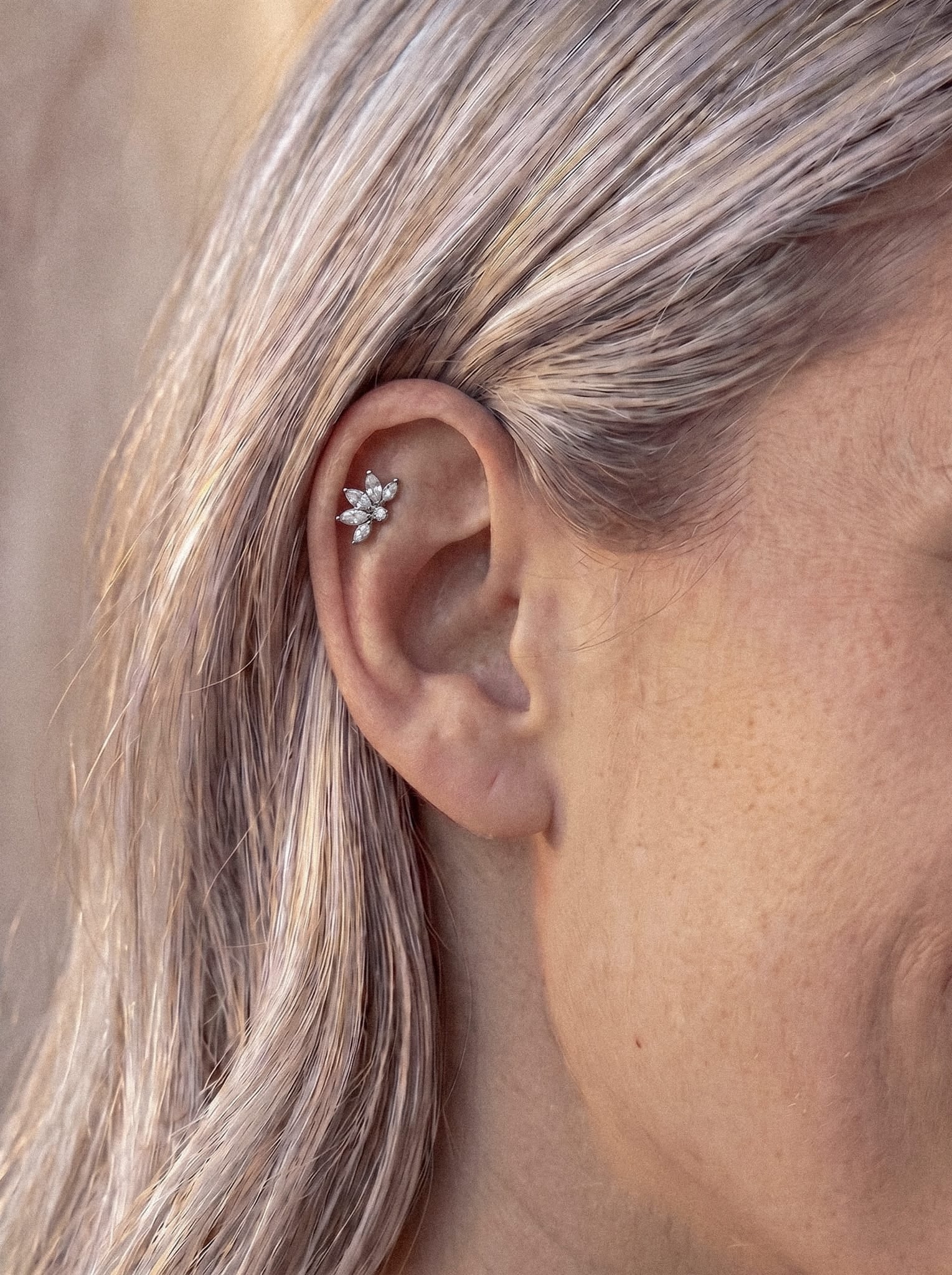 Un primer plano de la oreja de una persona adornada con el pendiente tipo «ear cuff» Ava de Titanio-Noble Art, una pieza con forma floral que incorpora cristales a modo de pétalos y que se lleva en la parte superior del cartílago. La persona tiene el pelo rubio claro y el fondo aparece ligeramente desenfocado.