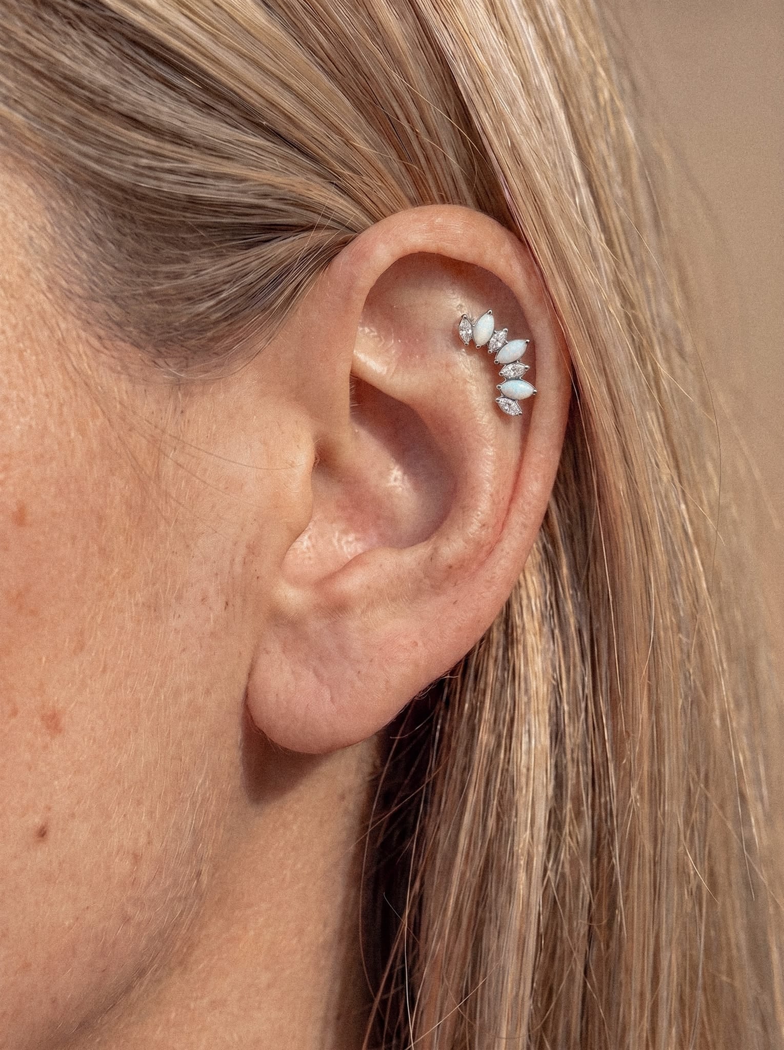 Primer plano de la oreja de una persona adornada con varios pendientes «Benus» de Titanio-Noble Art, con pequeñas piezas de plata en forma de marquesa a lo largo del cartílago. Varios pendientes lucen «ópalos» y «cristales» en forma de marquesa, lo que aporta al look un brillo celestial. La persona tiene el pelo liso y rubio claro, recogido detrás de la oreja.