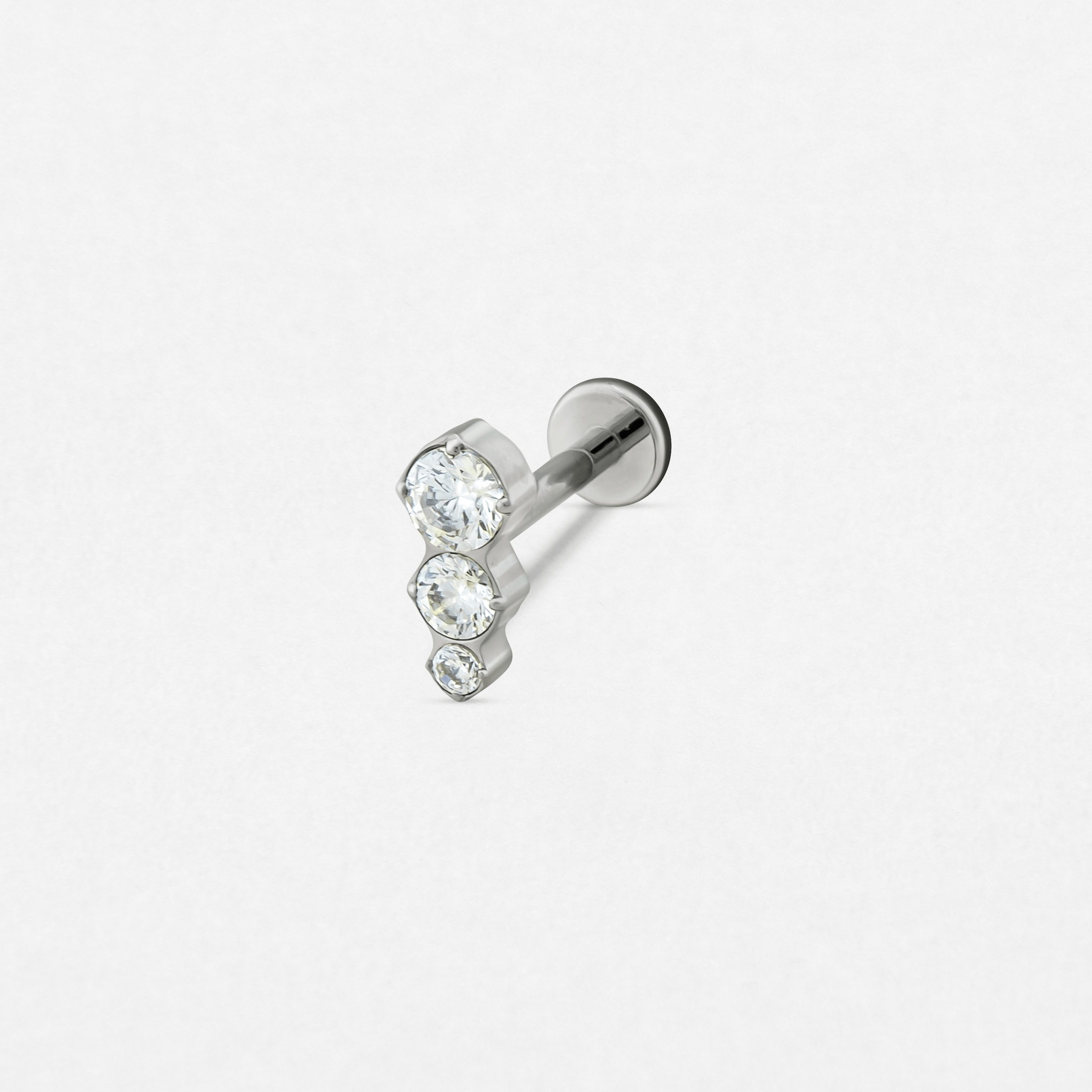 El pendiente de botón «Carles» de Titanio-Noble Art cuenta con tres piedras preciosas redondas y transparentes de distintos tamaños dispuestas verticalmente, ideales para realzar tu piercing. Se muestra sobre un fondo blanco liso.