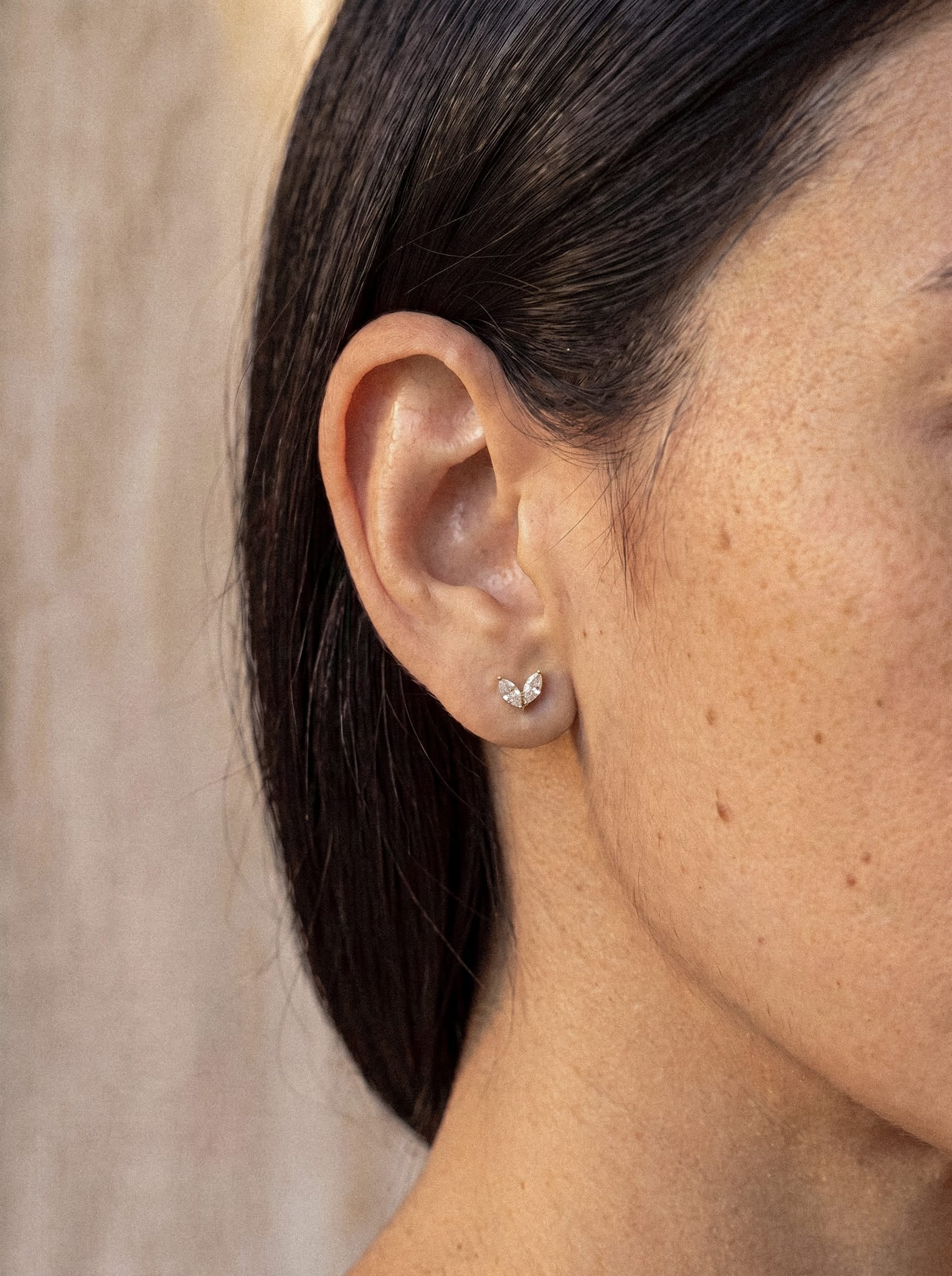 Un primer plano muestra la oreja de una persona adornada con el pendiente de botón «Creta» de Titanio-Noble Art: una pequeña pieza plateada con forma de ala cuyo delicado diseño, inspirado en la naturaleza, refleja la luz. El pelo liso y oscuro de quien lo lleva está cuidadosamente recogido detrás de la oreja, lo que resalta su piel suave y de tono marrón claro. Con una pared de color beige apagado como telón de fondo, esta joya de Titanio-Noble Art irradia una elegancia discreta.