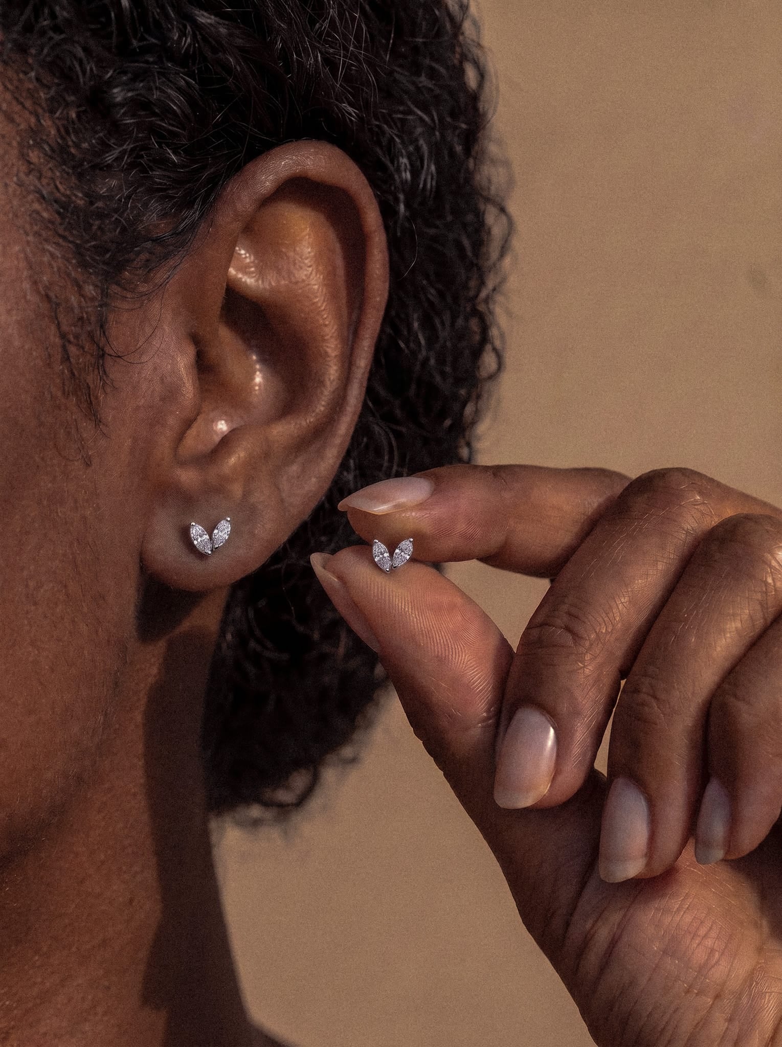 Una persona sostiene junto a la oreja un pequeño pendiente de botón plateado con forma de hoja de Creta, de la marca Titanio-Noble Art, mientras que en el lóbulo ya lleva puesto otro pendiente a juego. El fondo neutro centra la atención en los pendientes y en la oreja, resaltando la joyería inspirada en la naturaleza y el delicado diseño de este accesorio.