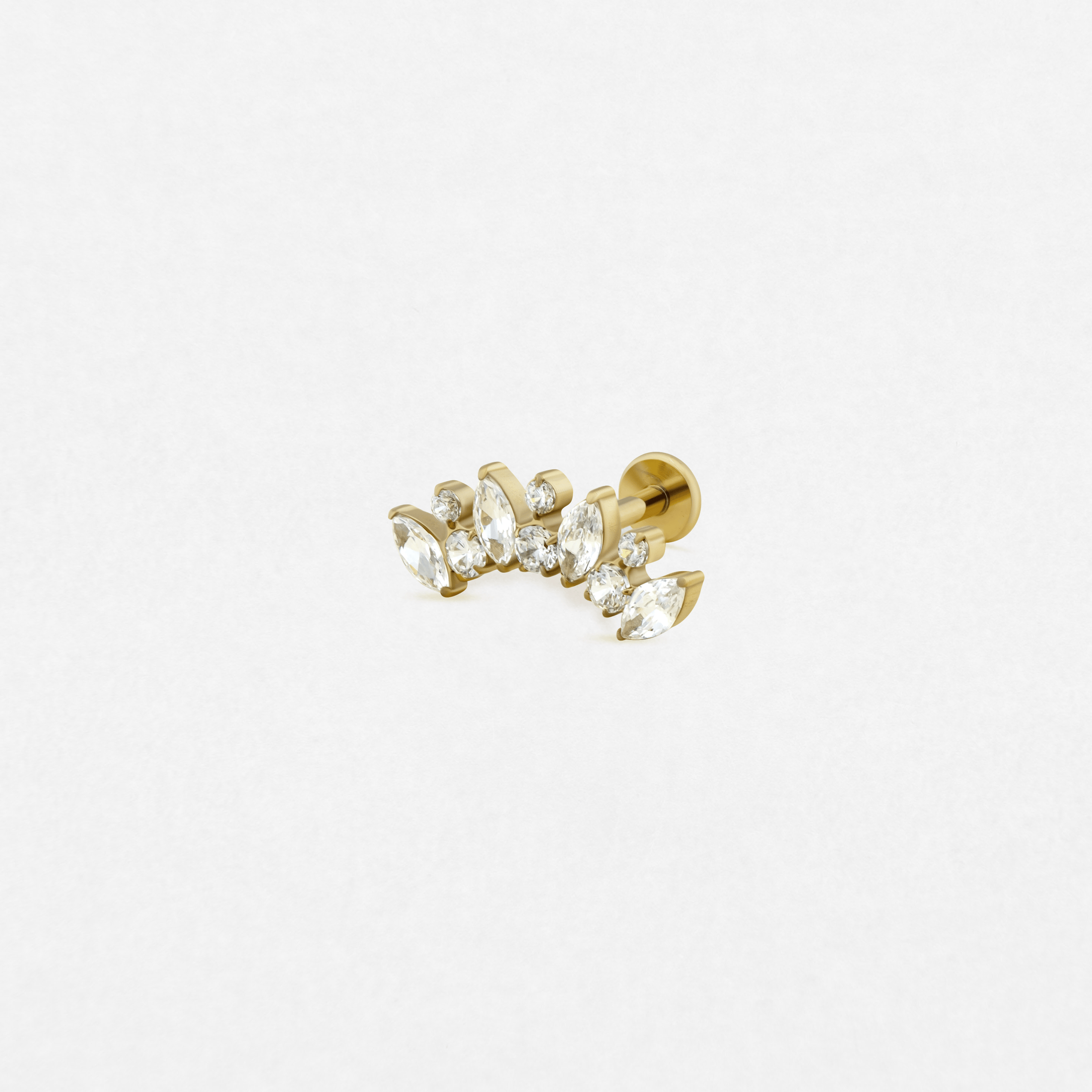 El modelo «Cluster Dario» de Titanio-Noble Art es un pendiente de botón dorado con piedras preciosas transparentes de corte marquesa y redondo, que destacan sobre un fondo blanco liso y brillan con elegancia, como una joya para la oreja en lugar de un tatuaje.