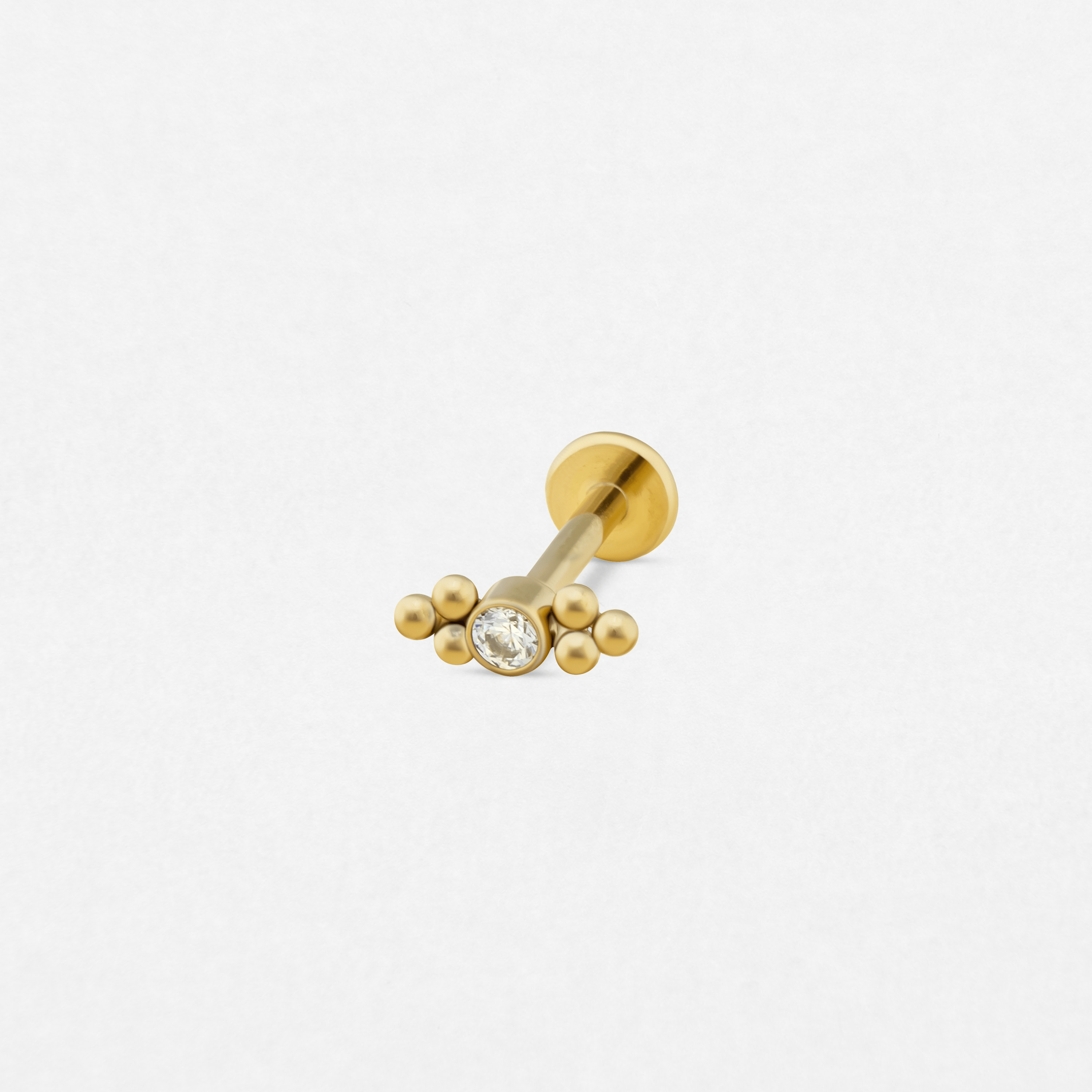 El pendiente «Destello doble» de Titanio-Noble Art cuenta con una piedra preciosa transparente en el centro, rodeada por debajo de cinco pequeñas cuentas doradas dispuestas en semicírculo; es ideal para piercings recientes y se muestra sobre un fondo blanco.