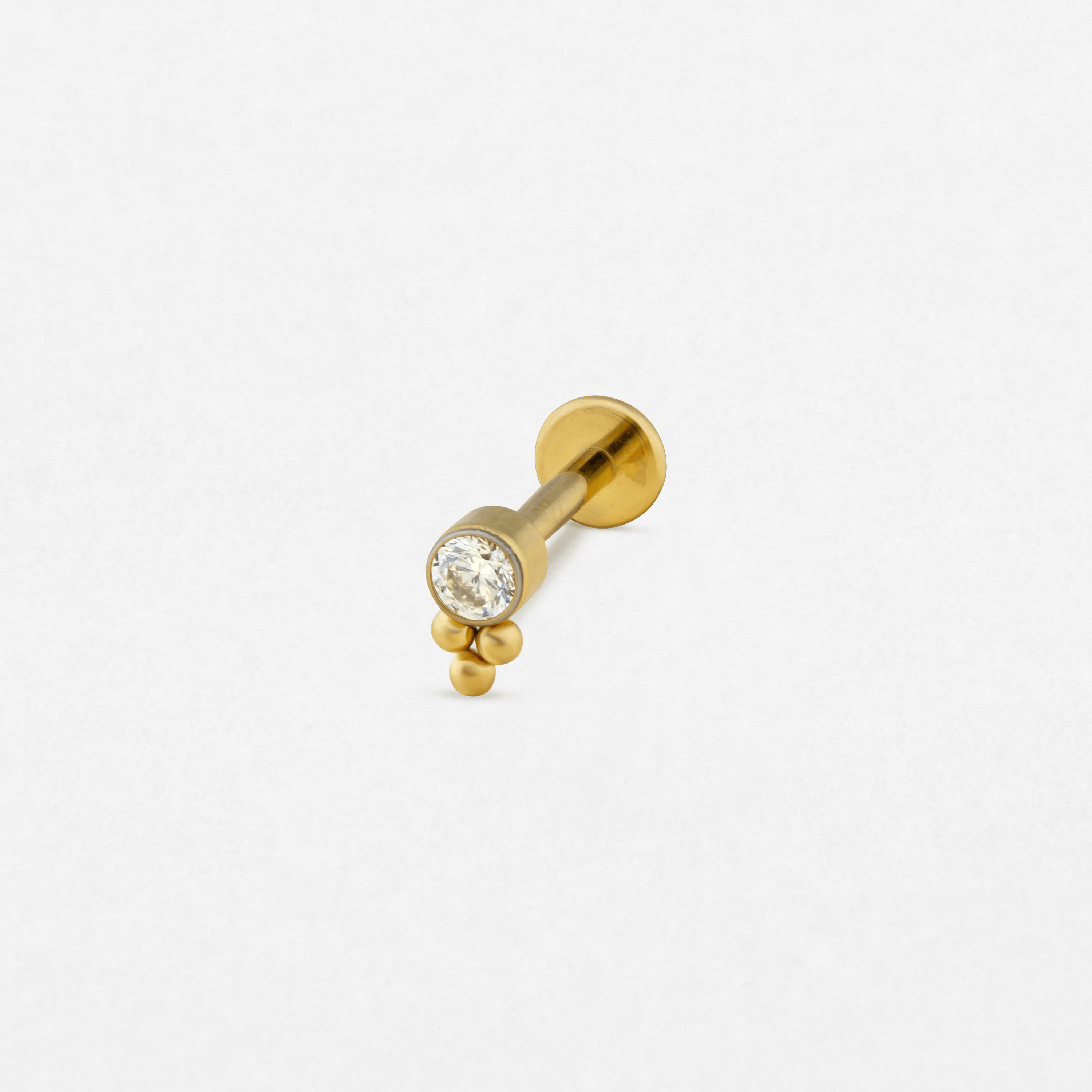 El modelo «Destello Simple» de Titanio-Noble Art es un pendiente de botón de oro con una gema redonda transparente y tres pequeñas cuentas de oro, ideal para cualquier tipo de piercing, que se muestra sobre un fondo blanco liso.